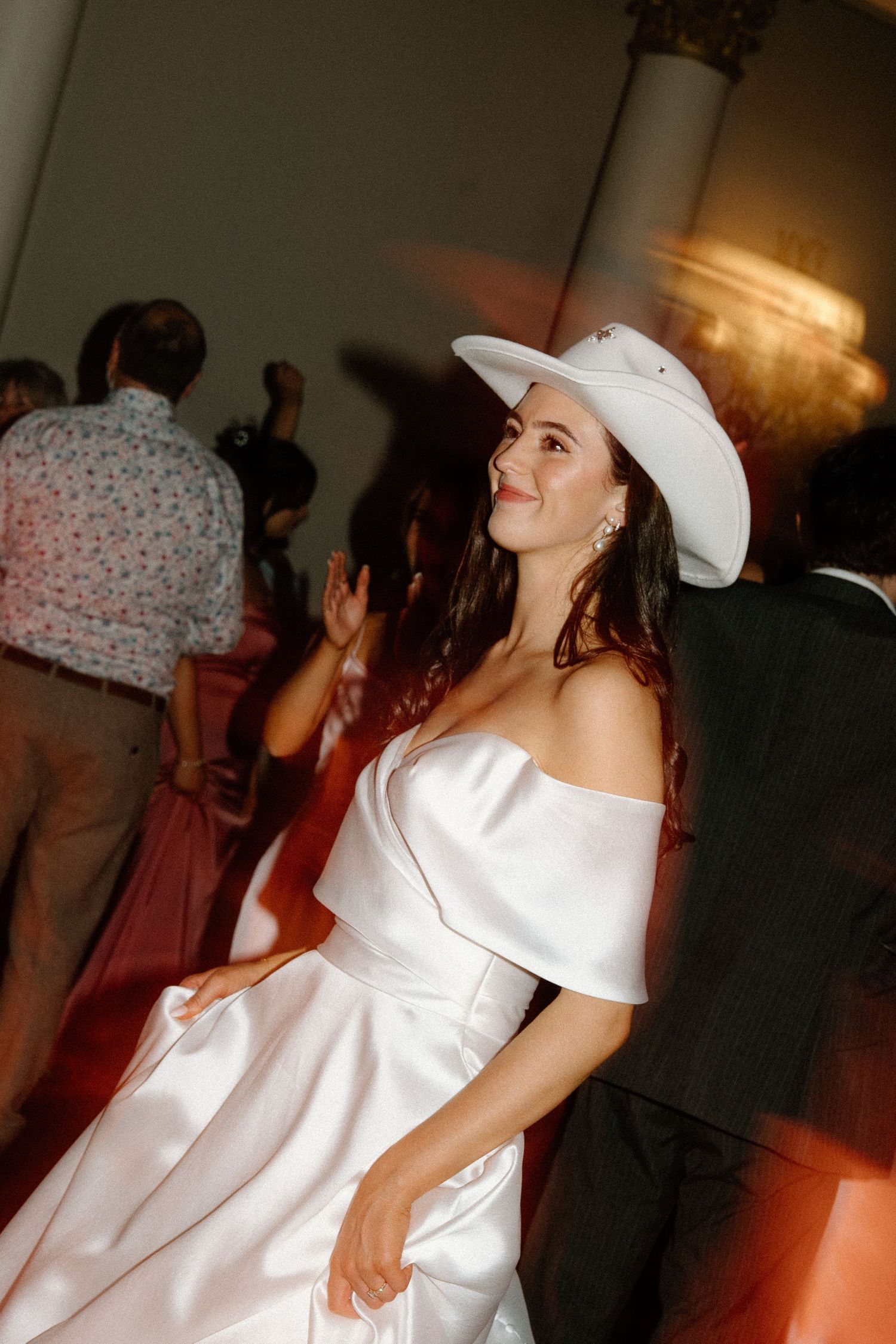 Bride dancing joyfully in white off-shoulder gown and white cowboy hat at wedding reception.