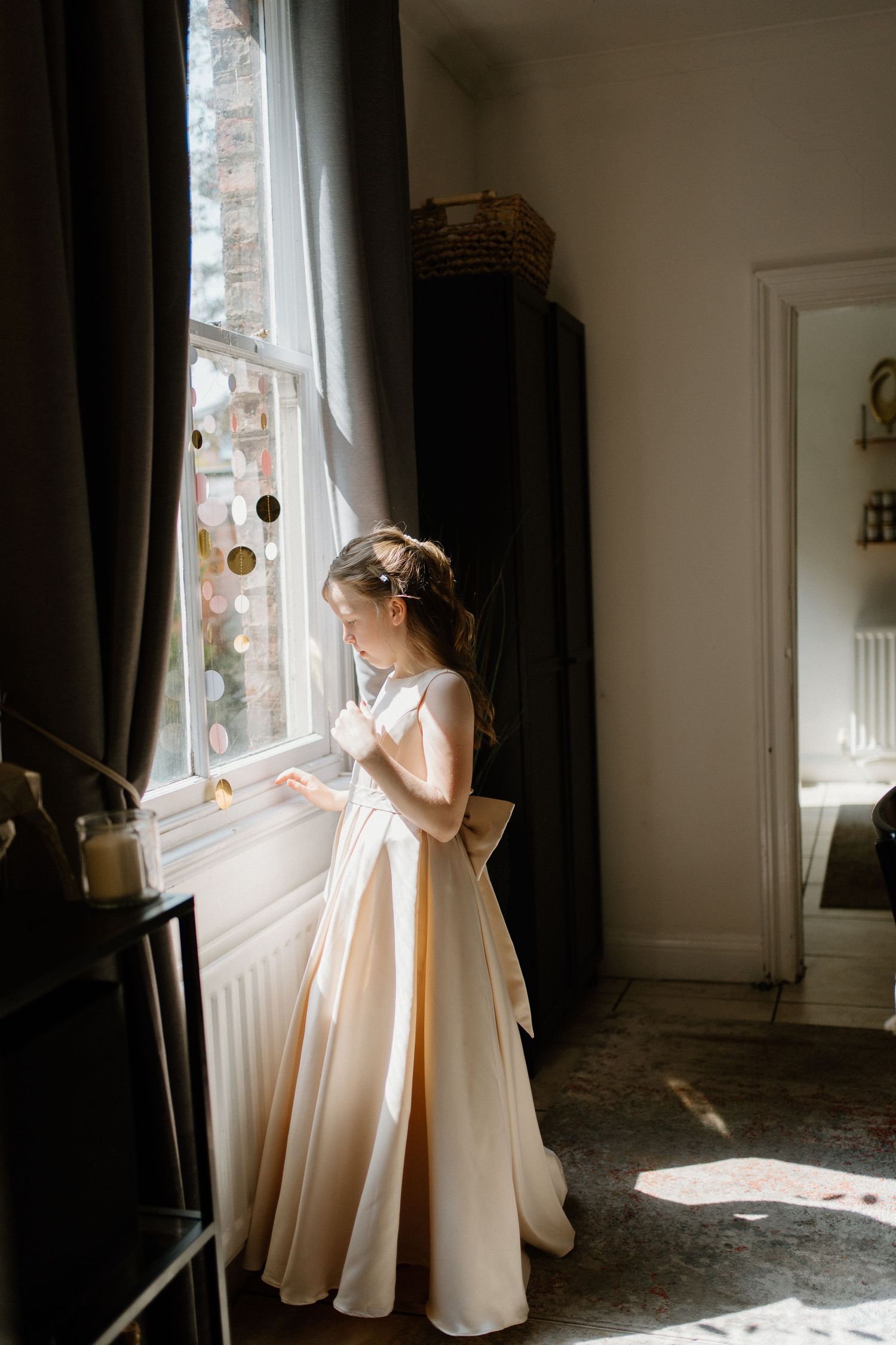 Elegant bride in off-shoulder satin gown with large bow standing by window in soft natural light.