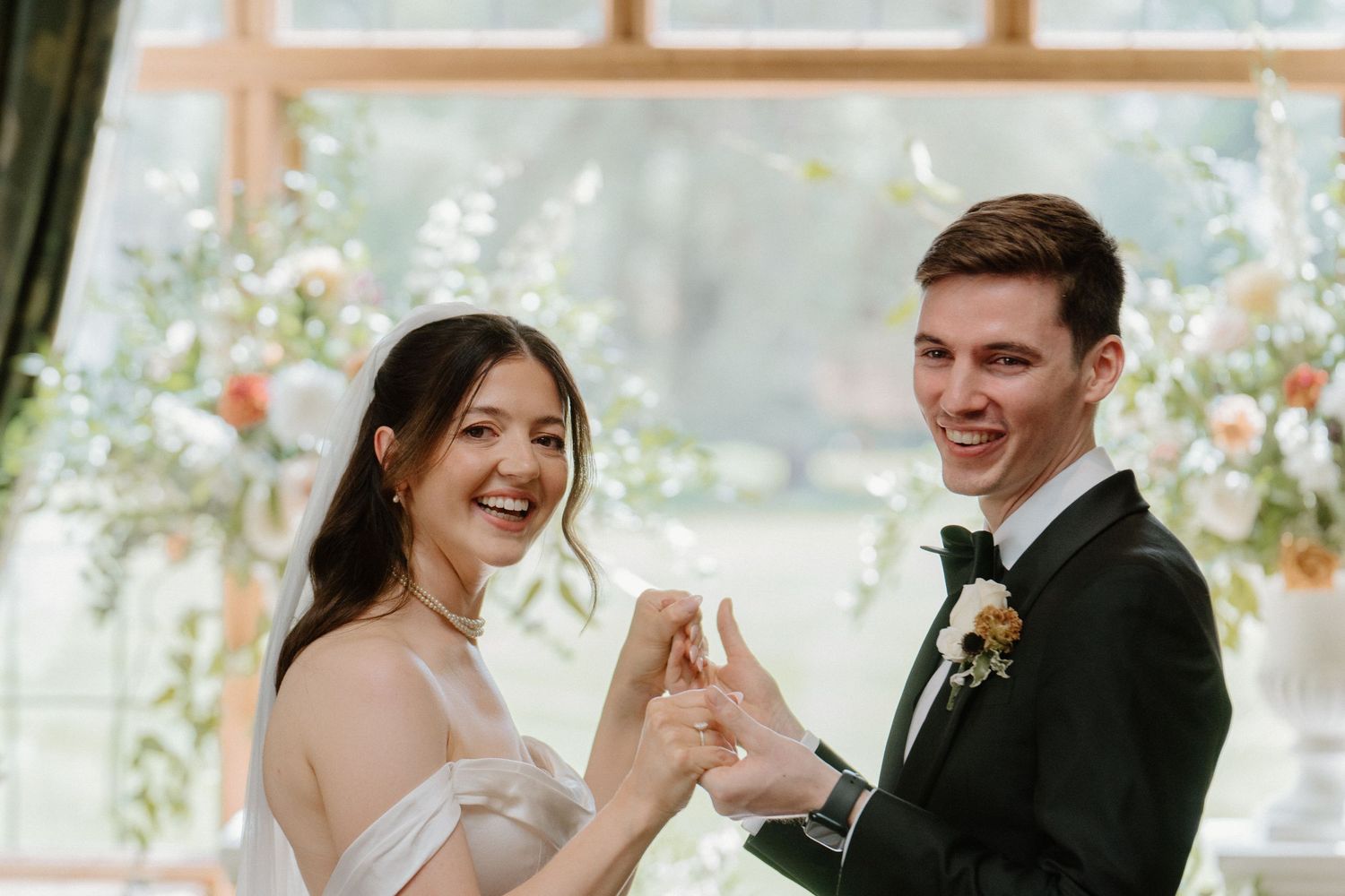Smiling bride and groom exchange rings during wedding ceremony with floral backdrop and natural light.