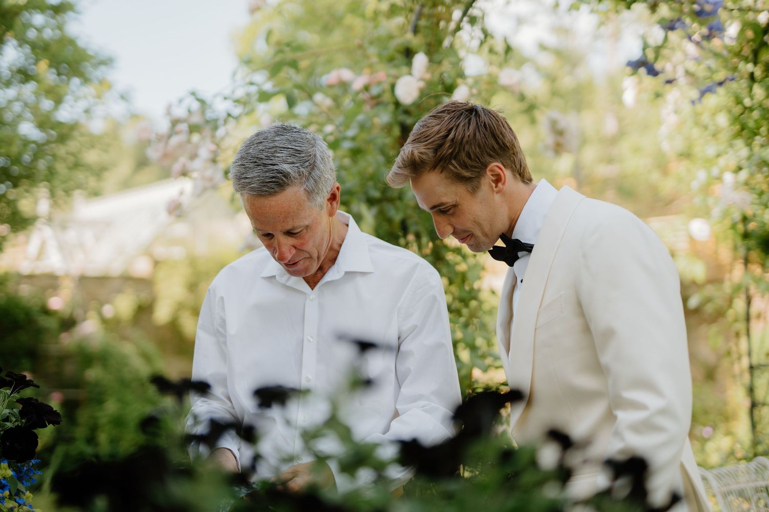 Groom in white tuxedo jacket shares a quiet moment with older man in garden wedding setting.