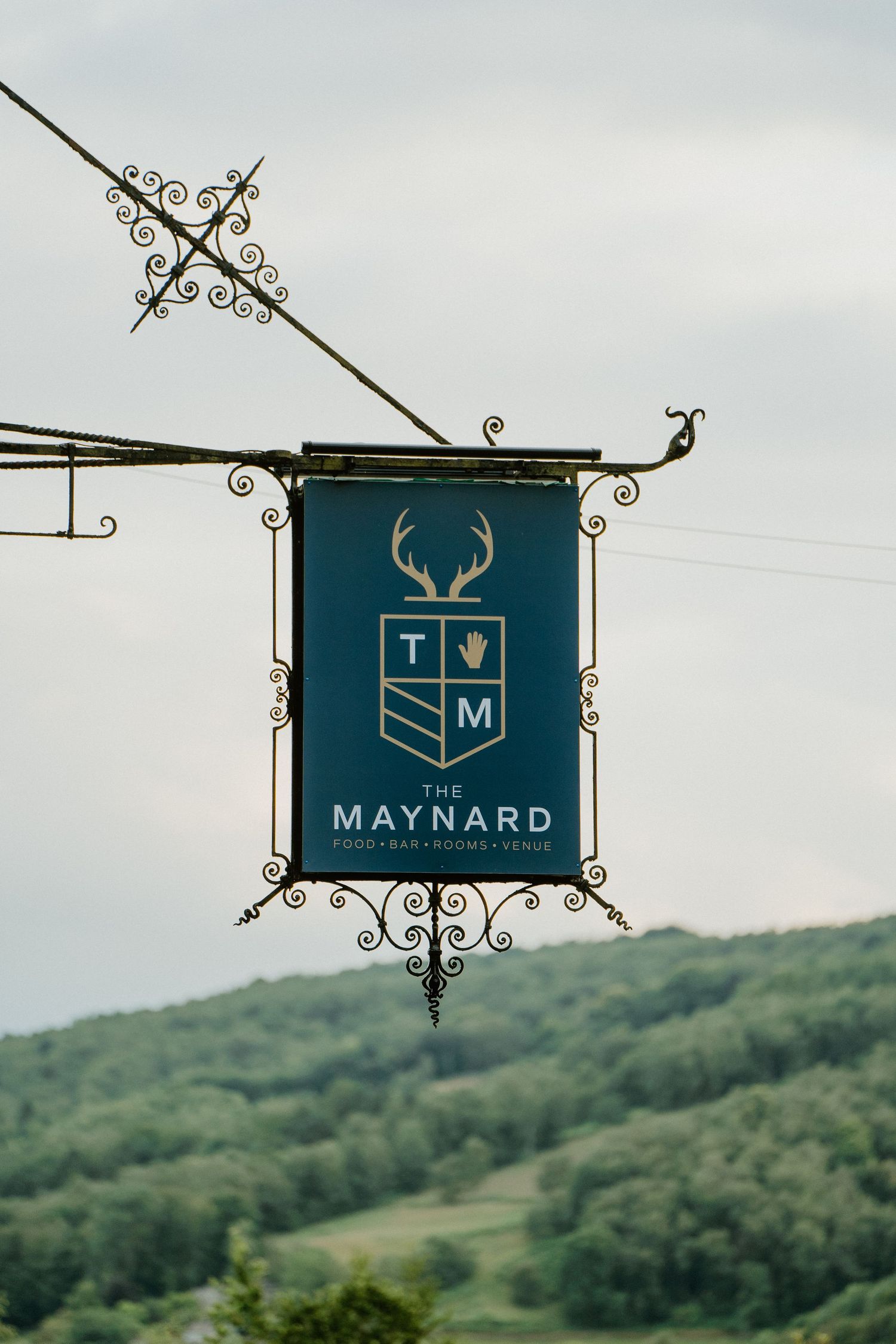 The Maynard wedding venue sign with decorative ironwork against green hillside backdrop.
