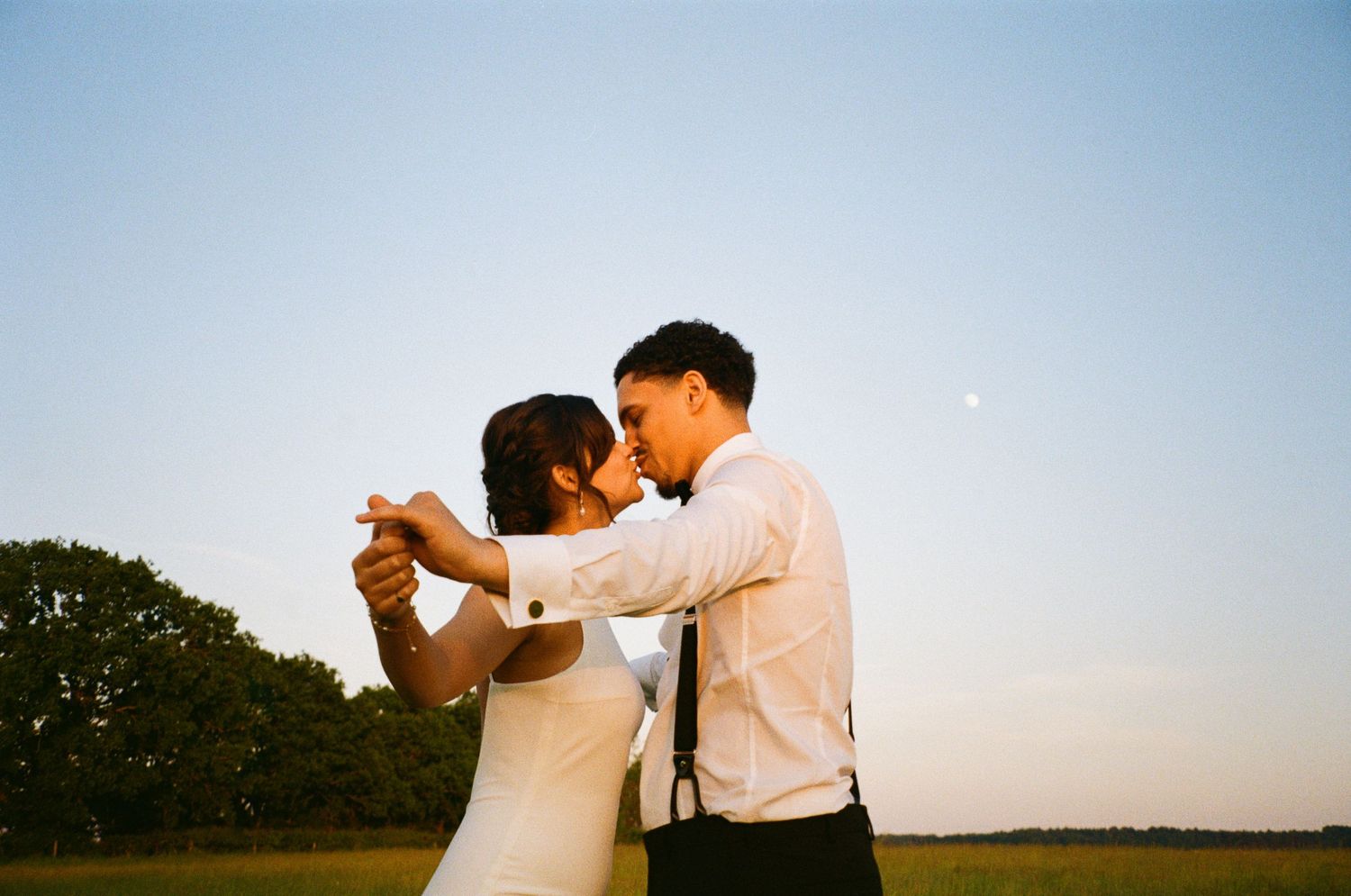 Bride and groom sharing a romantic kiss in an open field during golden hour sunset.