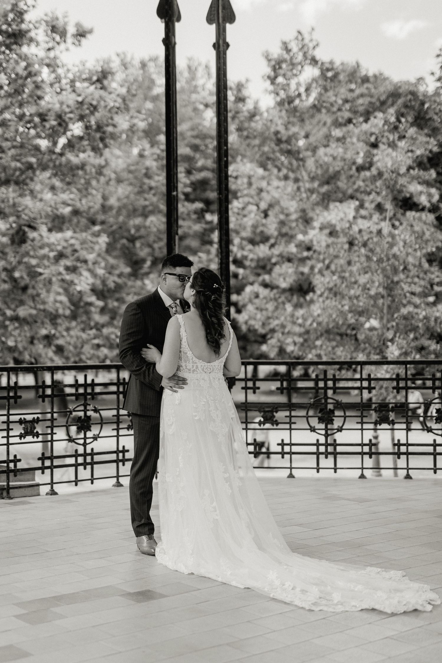 Black and white photo of bride and groom sharing a kiss on an outdoor terrace with iron railings and trees.