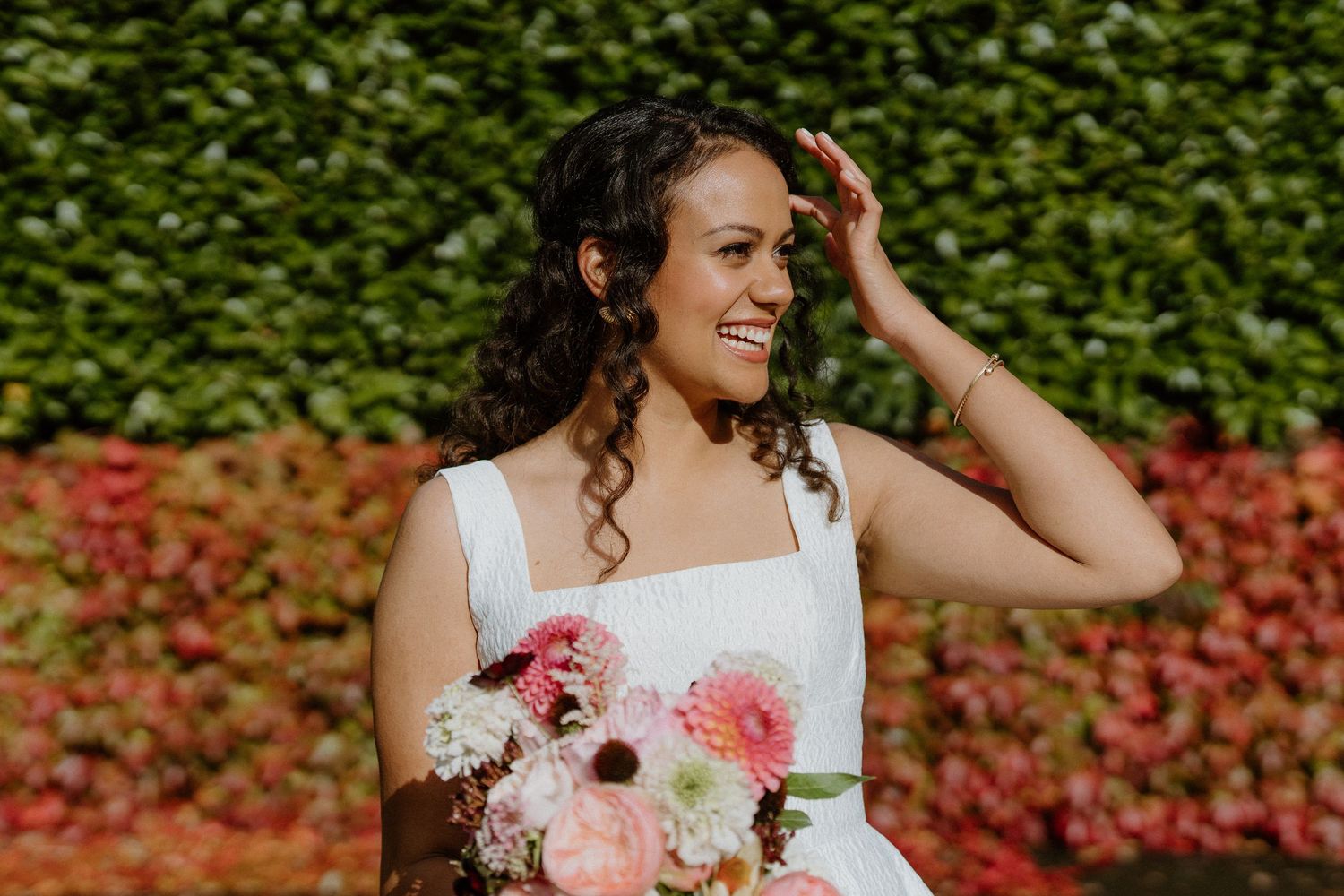Smiling bride holding pink bouquet in front of colorful foliage wall in outdoor wedding portrait.