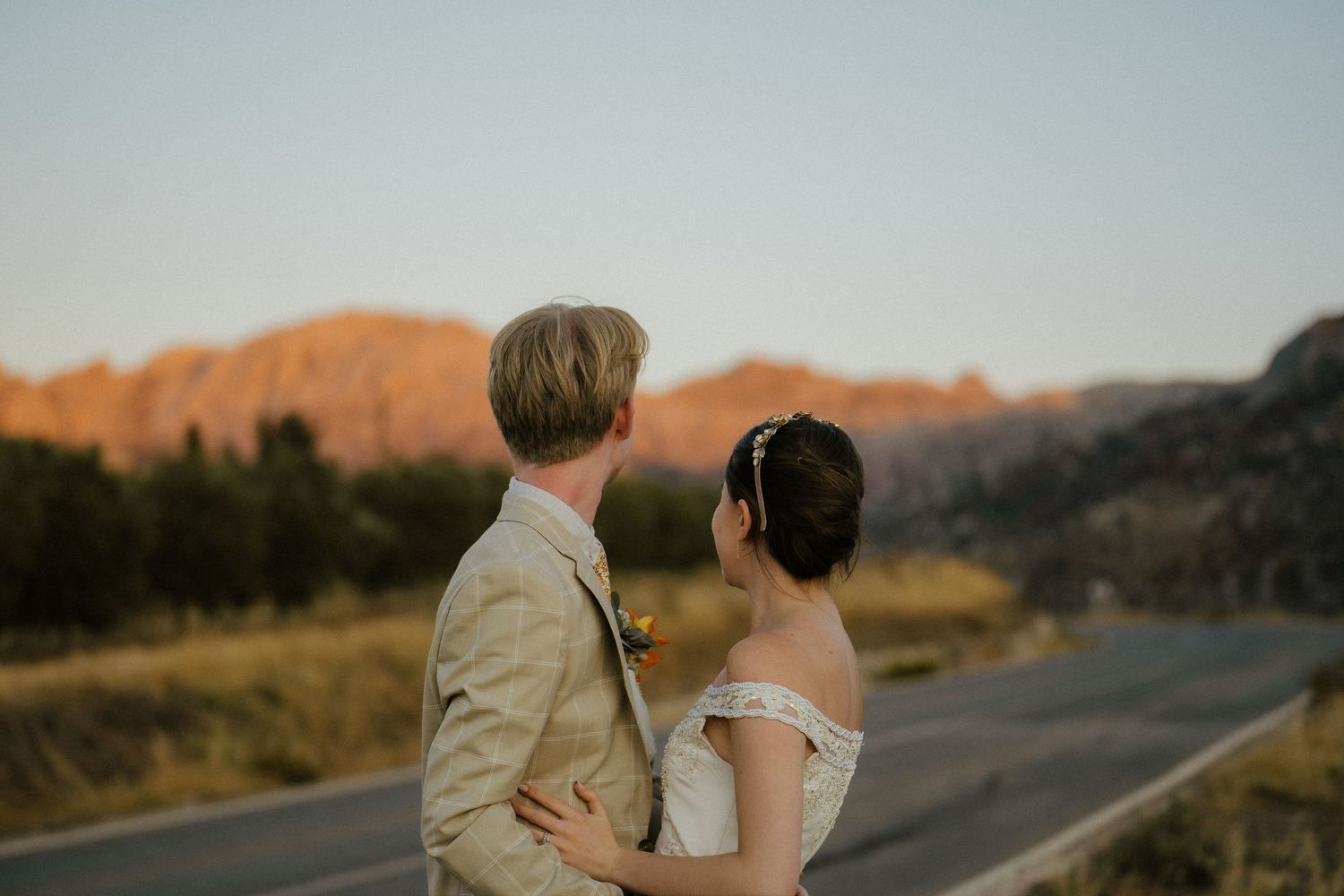 Bride and groom embrace on a scenic road with red rock mountains glowing at sunset in the background.