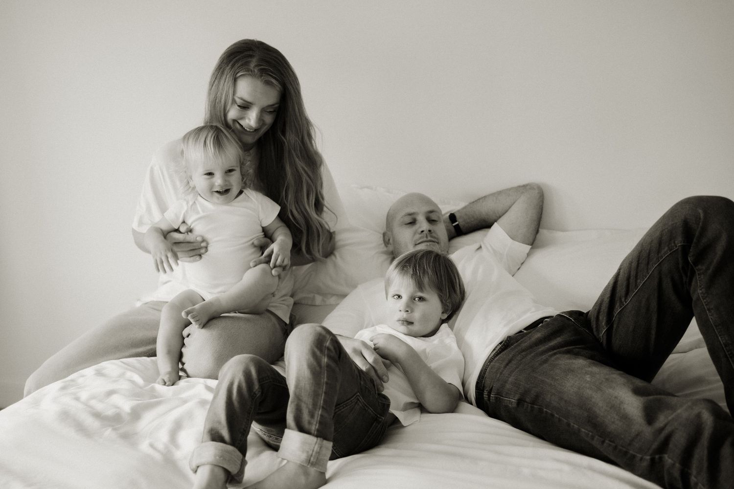 Black and white family photo of parents and two young children relaxing together on a white bed.
