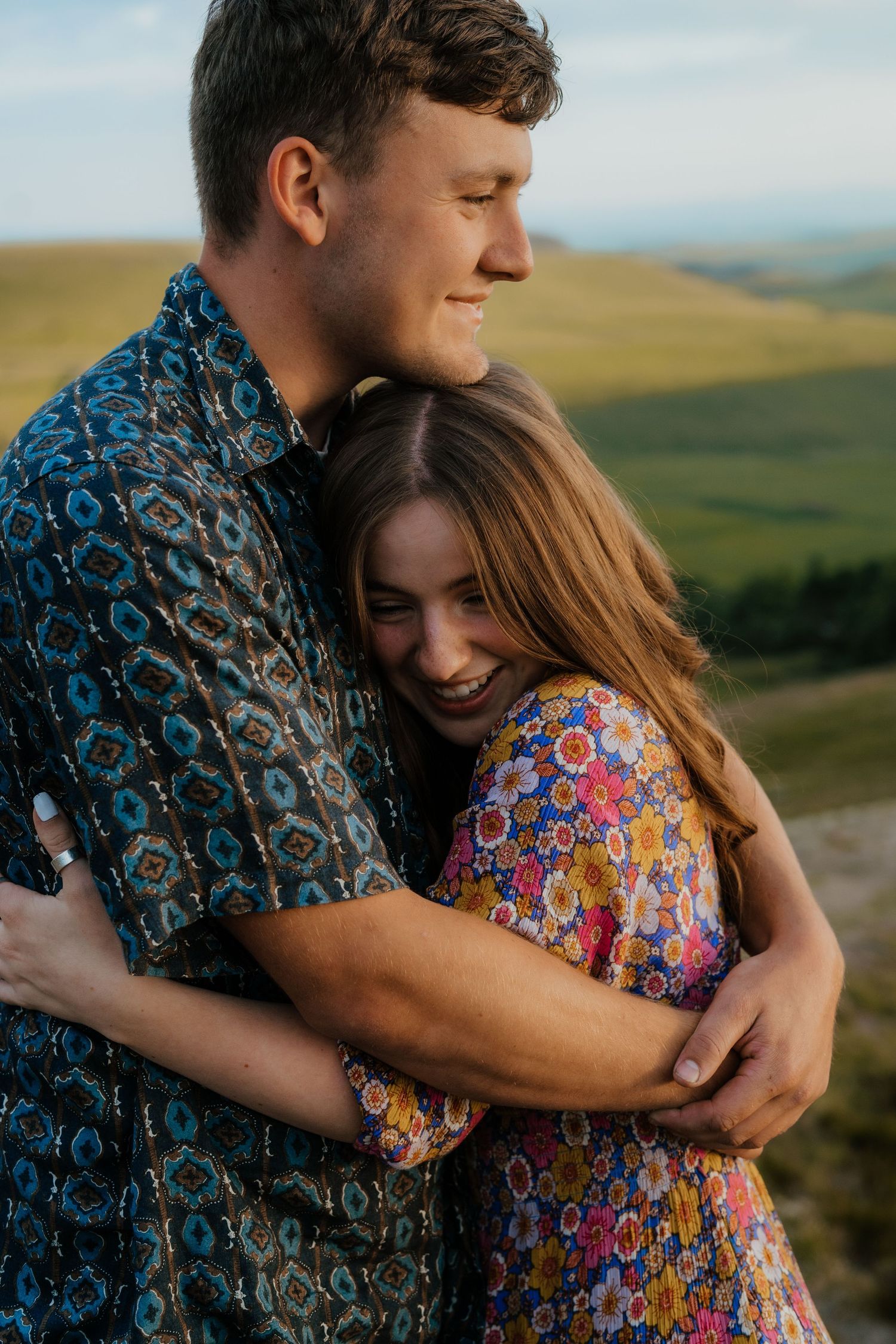 Laughing woman hugging her smiling partner on a scenic green hillside at sunset.