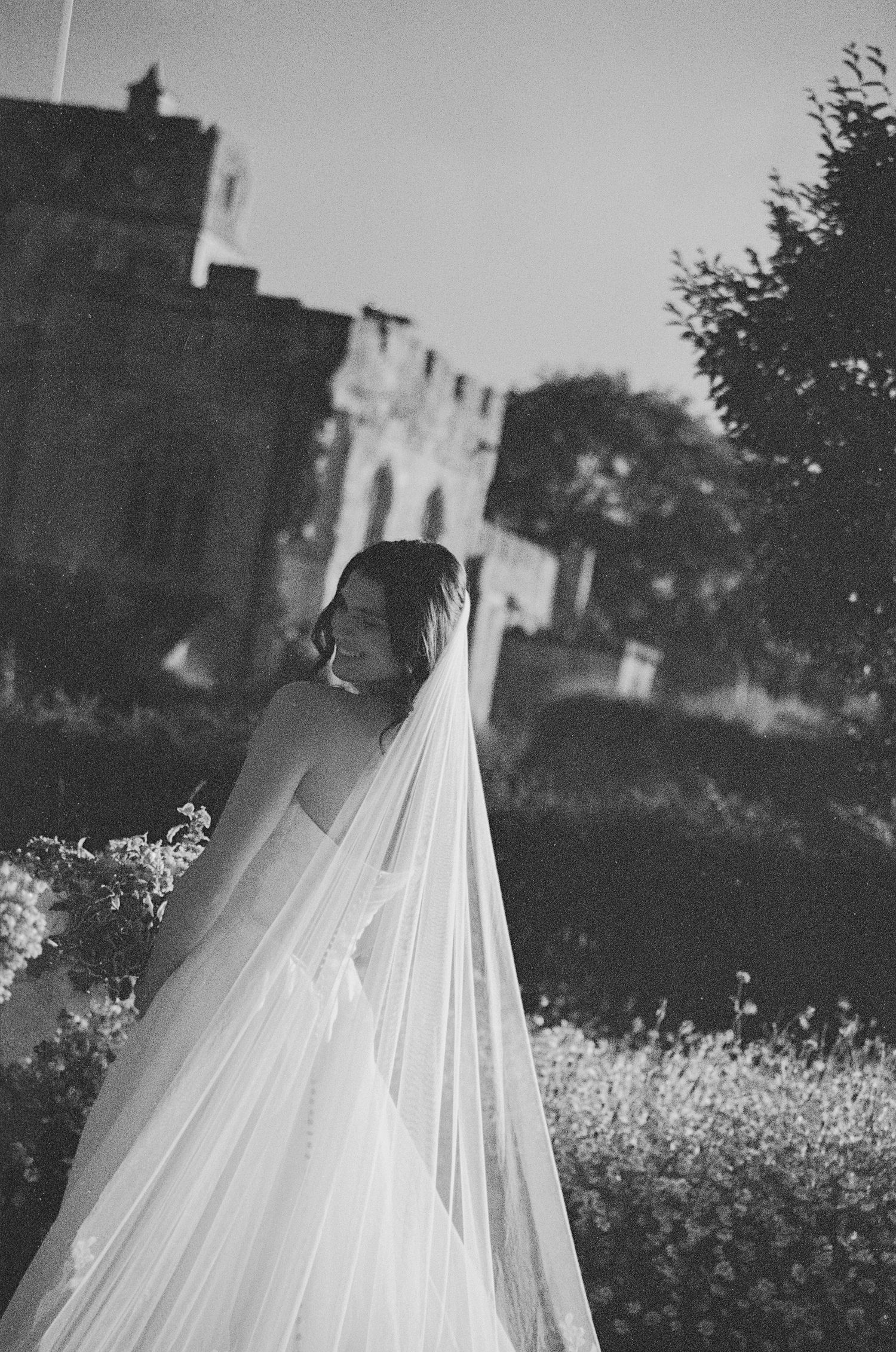 Black and white portrait of a smiling bride in a flowing veil outside a historic castle venue.