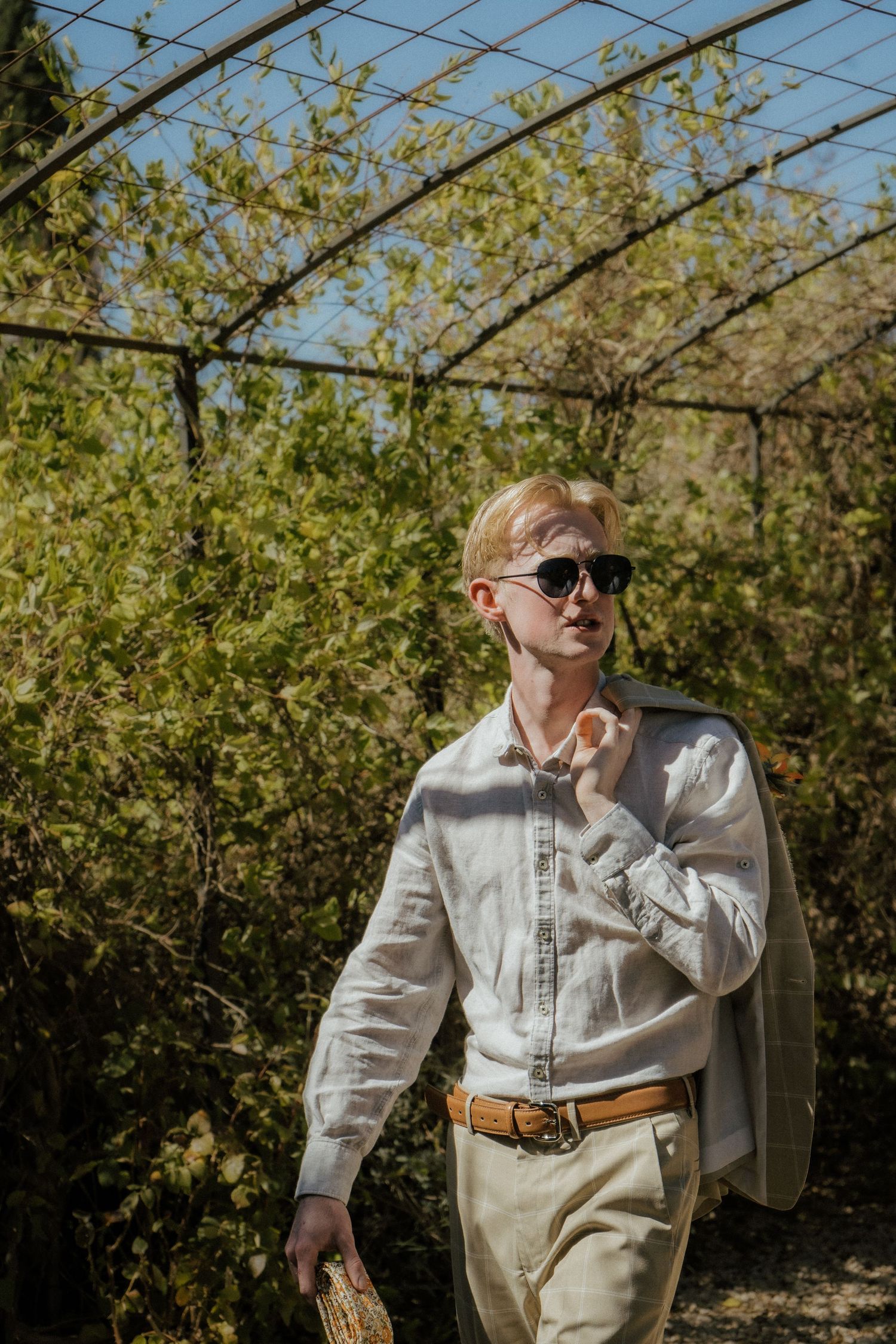 Blonde groom in linen shirt and sunglasses walking through garden pergola on sunny day.