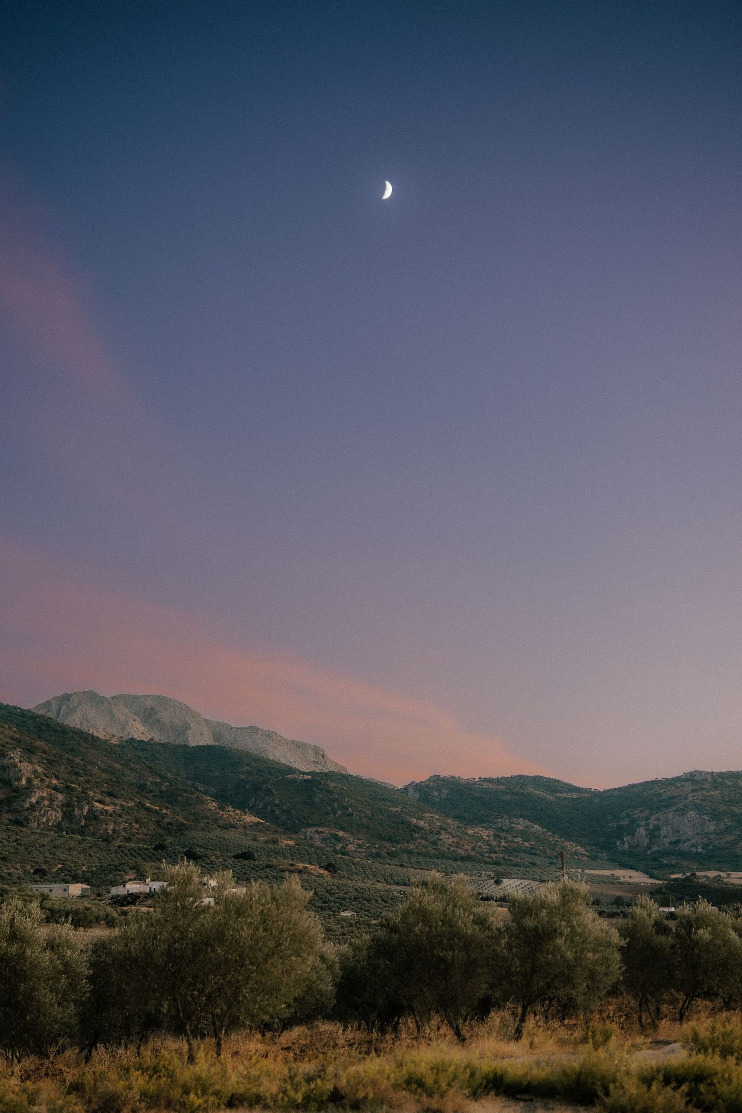 Crescent moon rising over scenic mountain landscape at twilight with pink and purple sky.