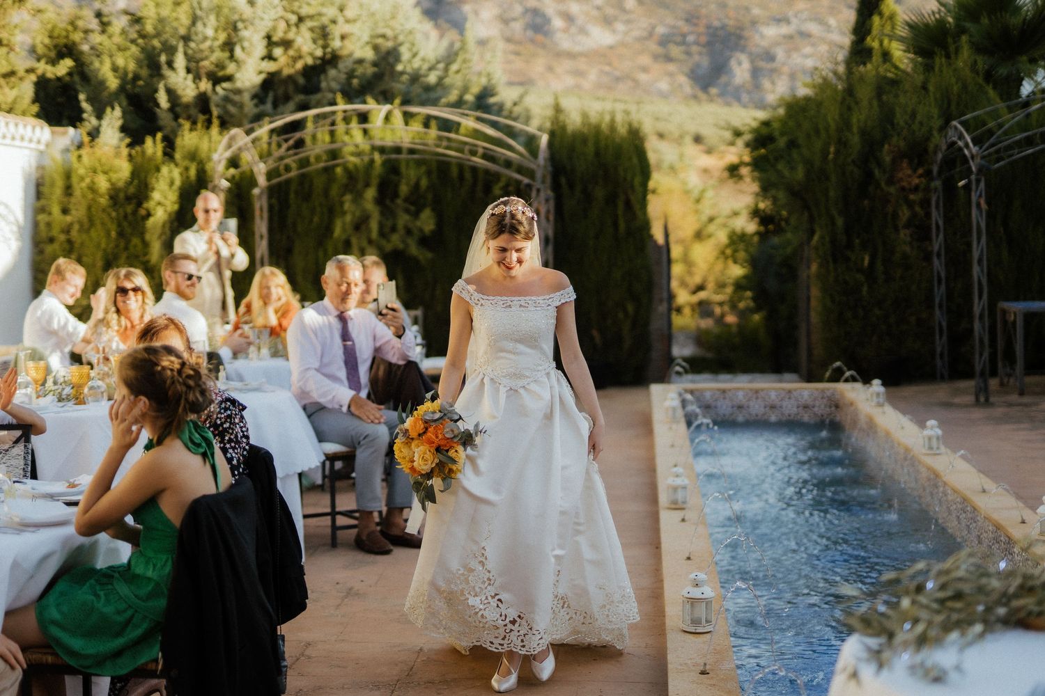 Bride in white gown carrying yellow bouquet walking beside pool at outdoor wedding reception.