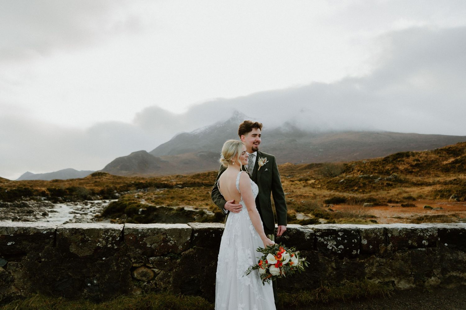 Bride and groom stand on a stone bridge surrounded by misty Scottish Highland mountains.