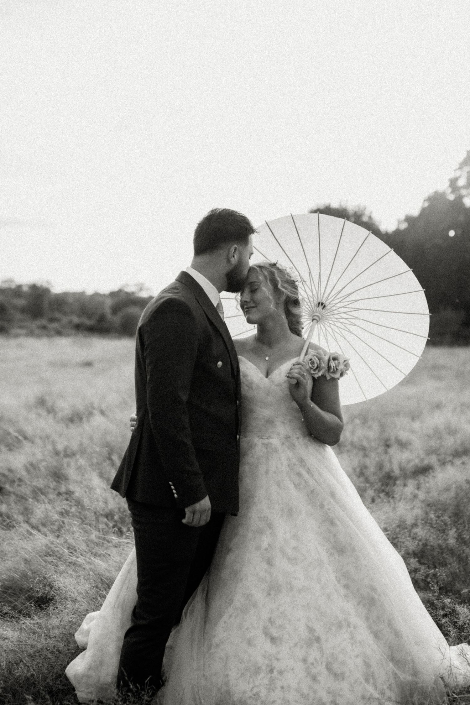 Black and white photo of bride and groom sharing intimate moment in field, bride holding parasol.