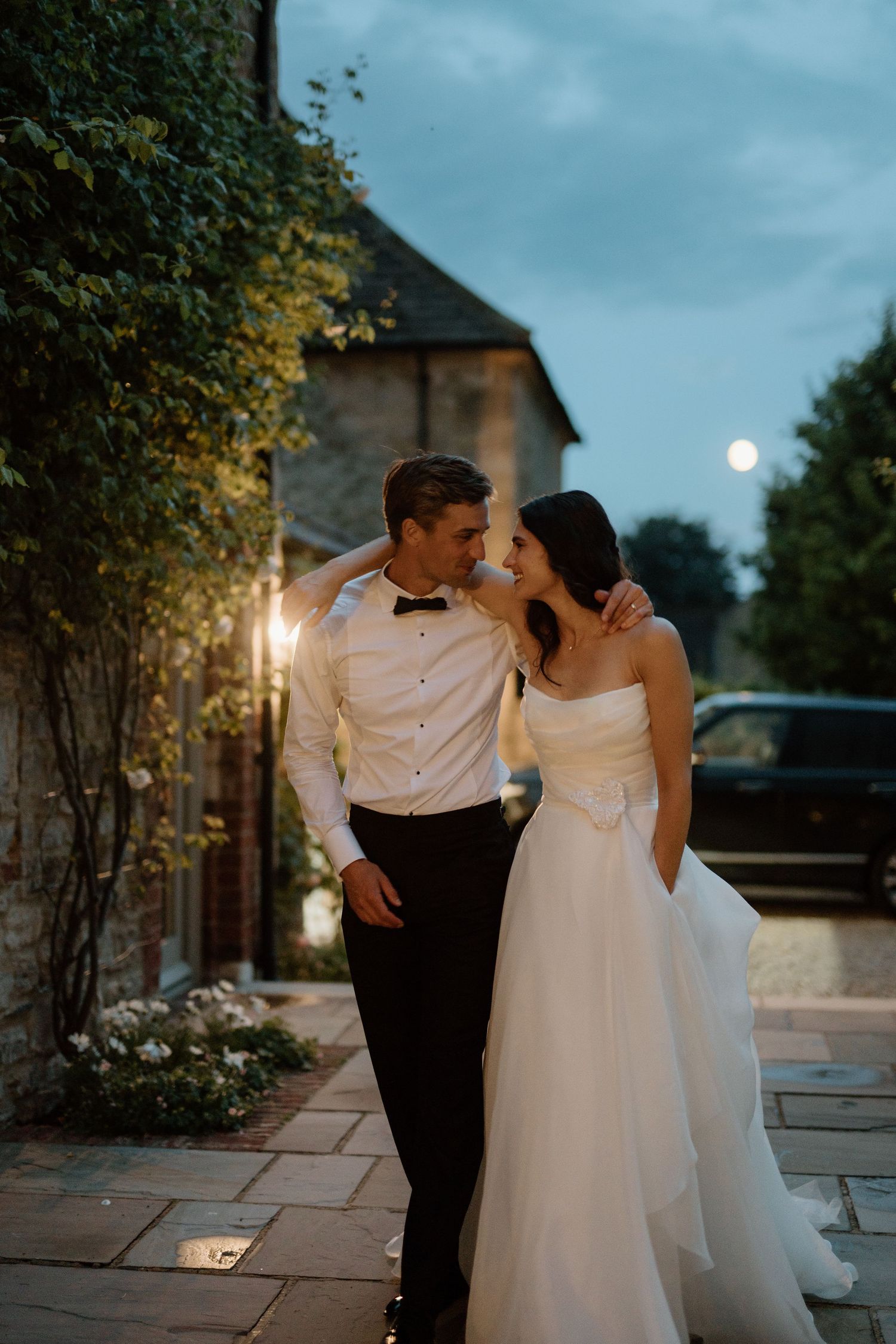 Bride and groom sharing romantic moment outdoors at dusk near stone building.