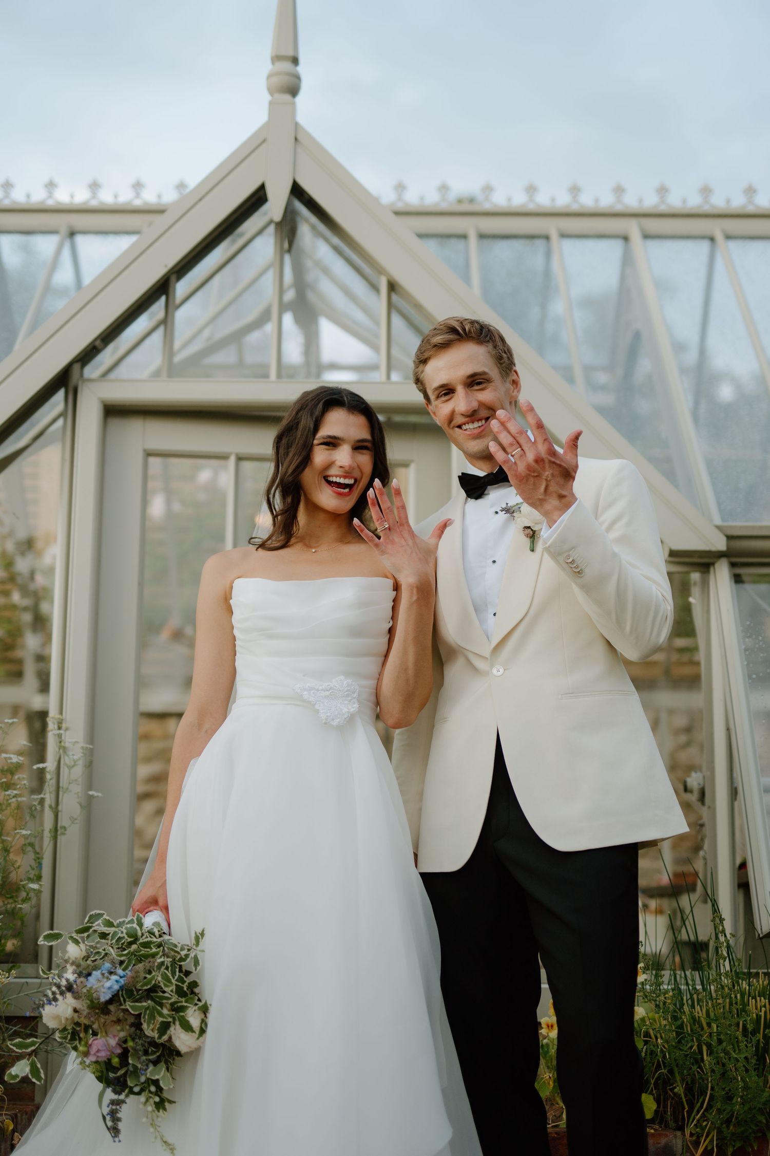Smiling bride and groom show off wedding rings outside a glass greenhouse, groom in white tuxedo jacket.