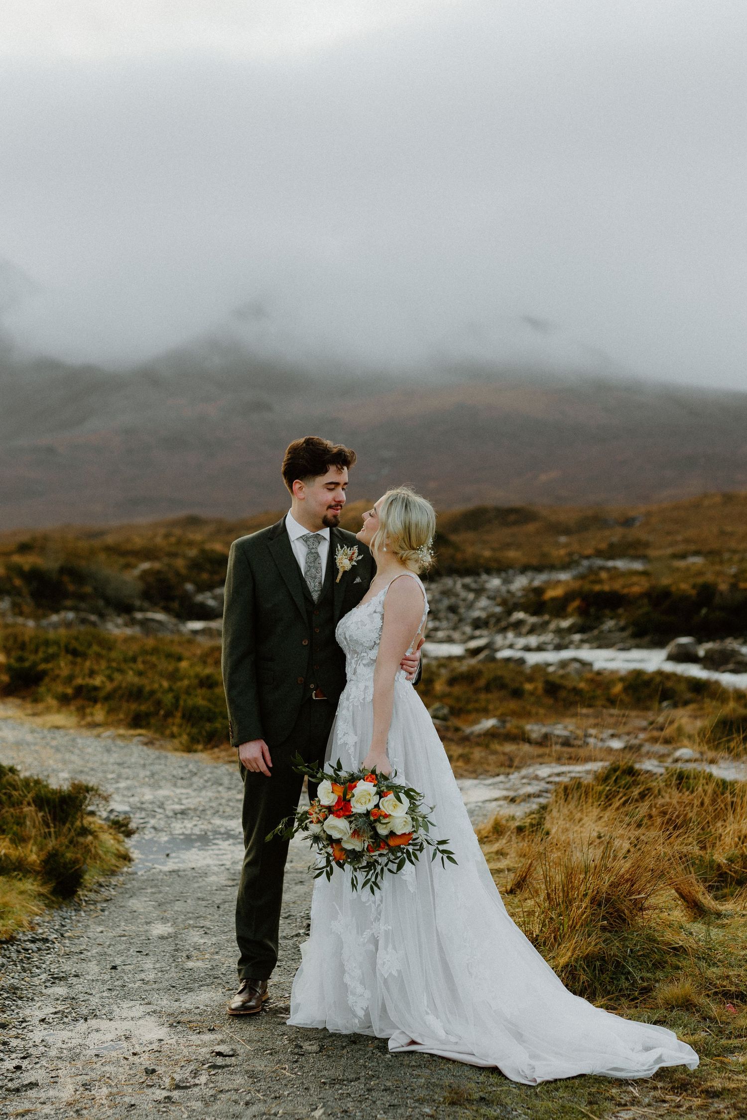 Bride and groom share intimate moment on misty Highland gravel path, bride holding colorful bouquet of roses.