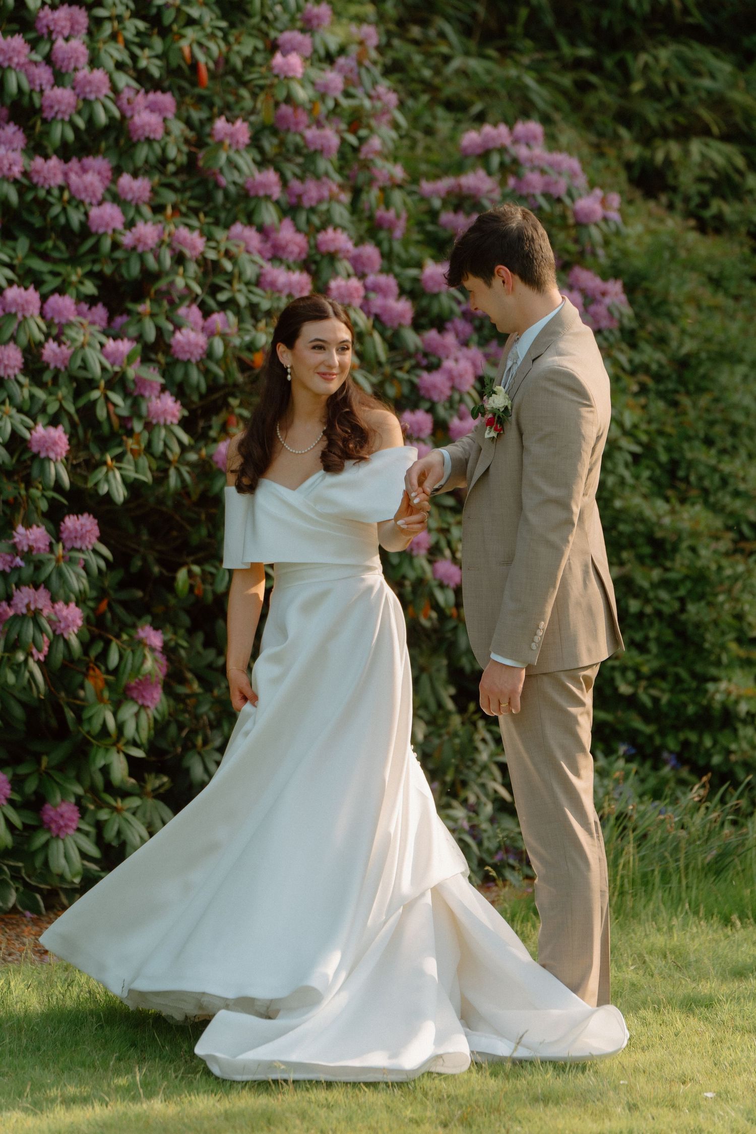 Bride twirling white gown while groom watches beside lush purple flowering rhododendron bushes.