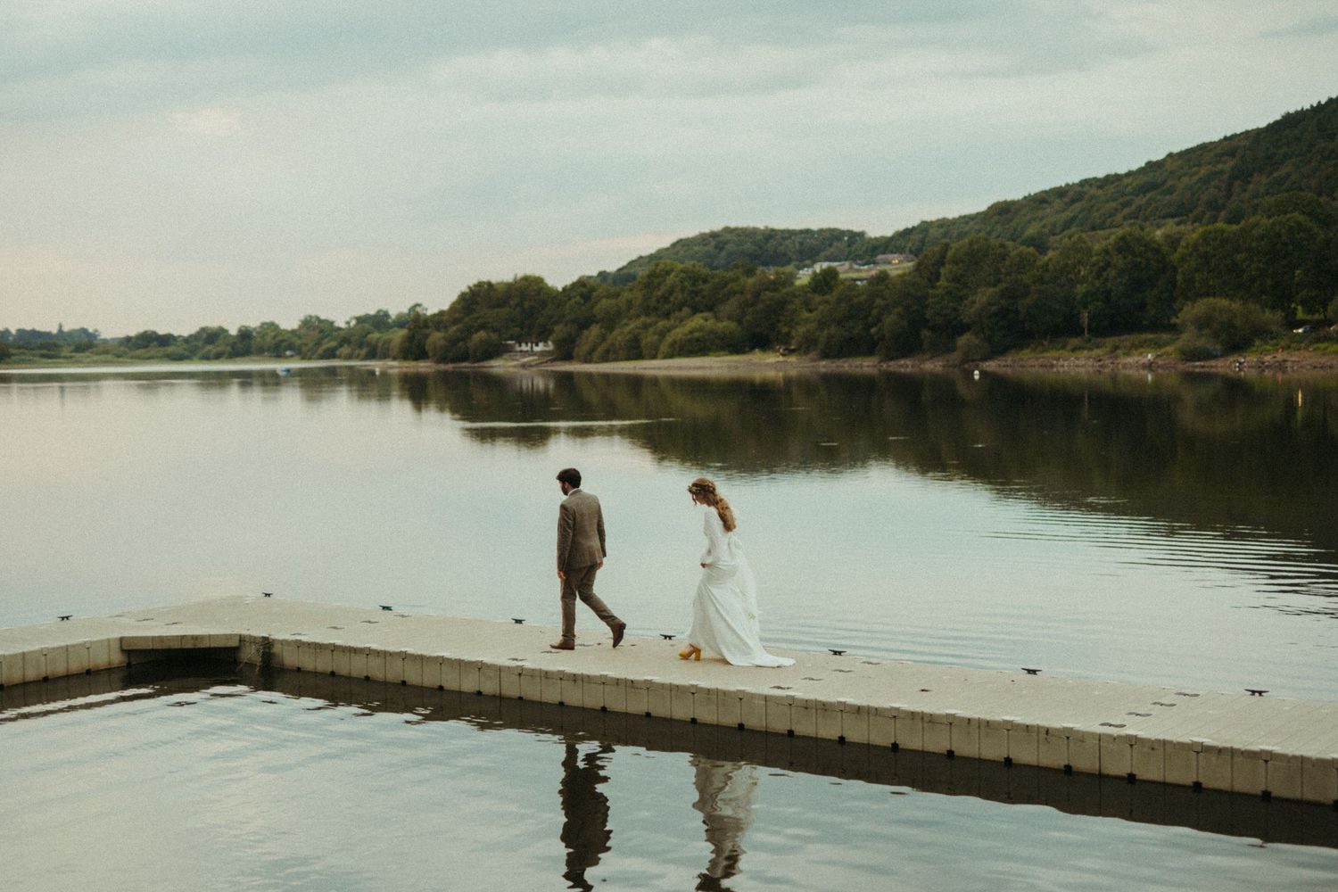Bride and groom walking hand in hand on a lakeside dock with calm water and green hills in the background.