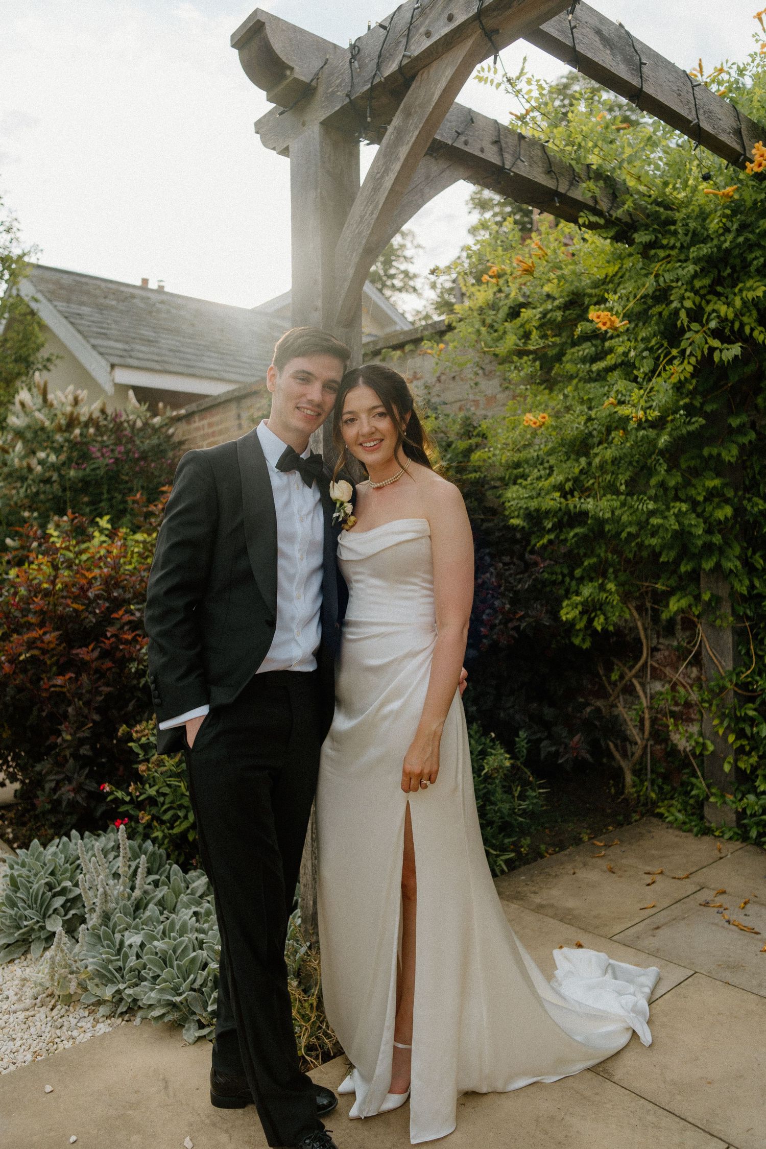 Bride in white satin gown and groom in black tuxedo posing under a wooden pergola.