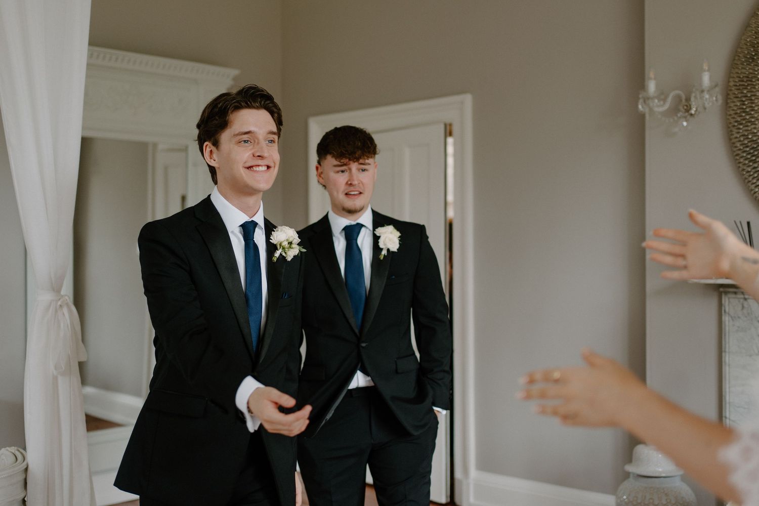 Two groomsmen in black suits with white boutonnieres smiling in a bright elegant wedding venue room.