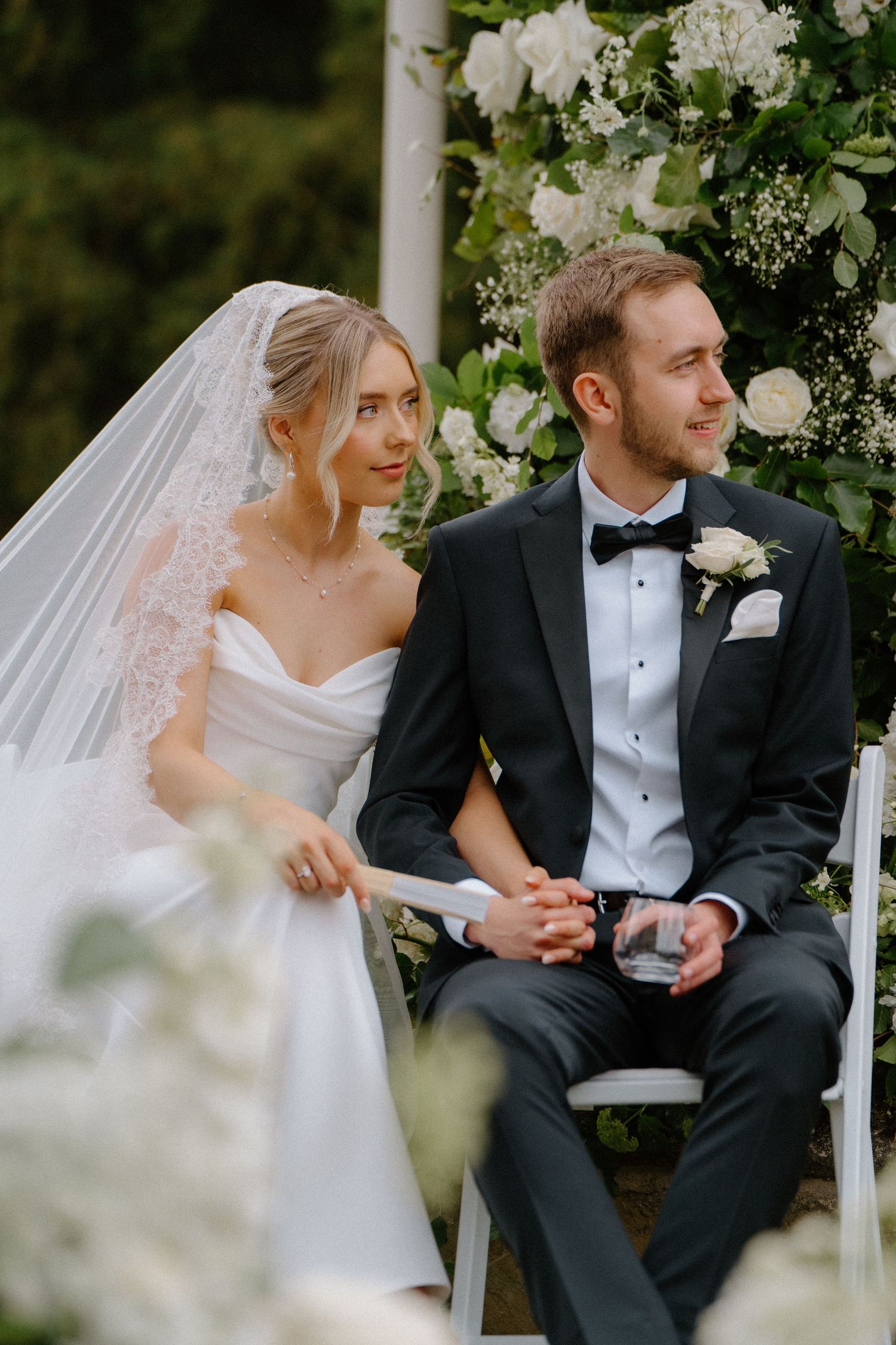 Bride and groom sit together at outdoor altar adorned with white floral arch during wedding ceremony.