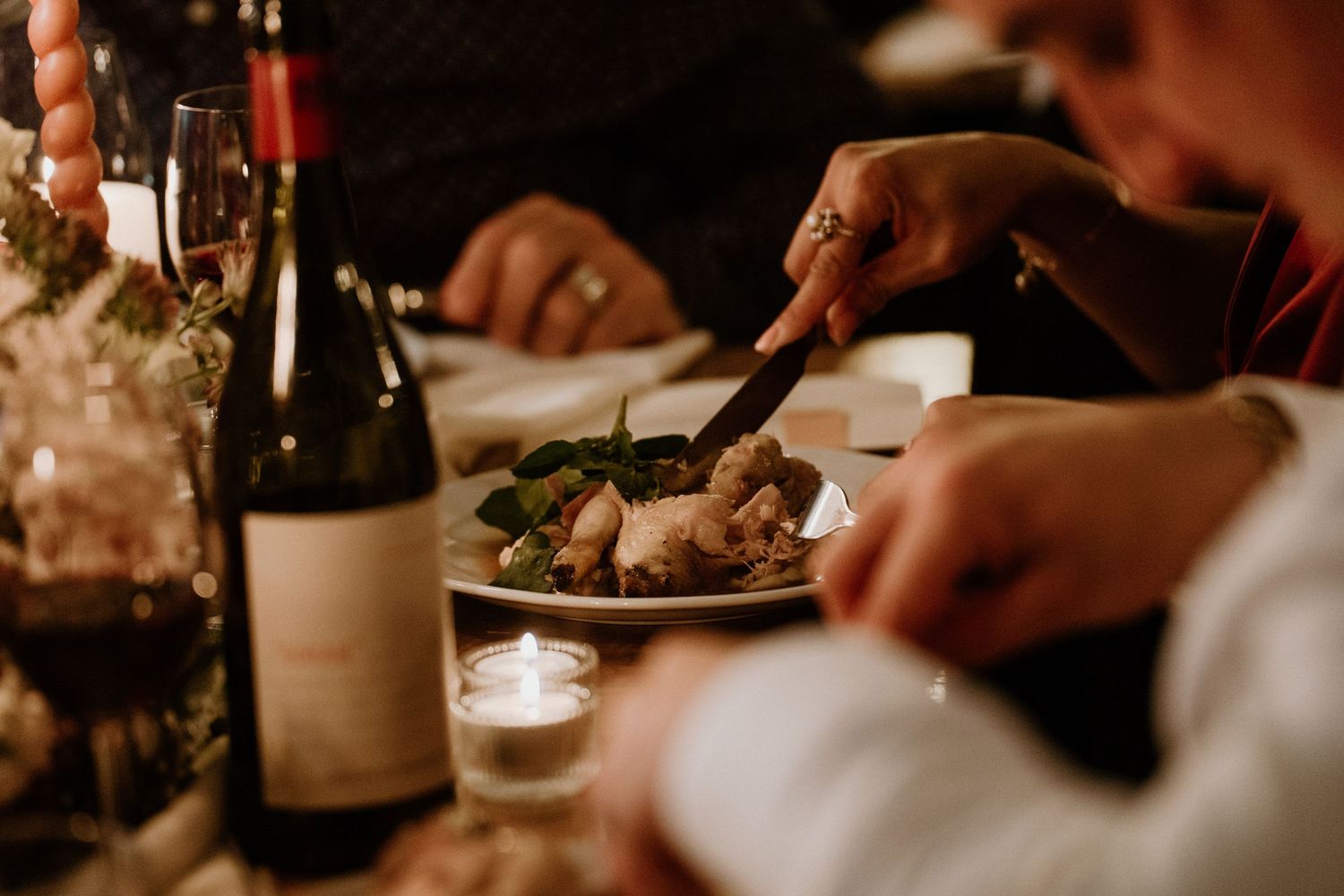 Close-up of wedding guest cutting food at a candlelit reception table with wine and florals.