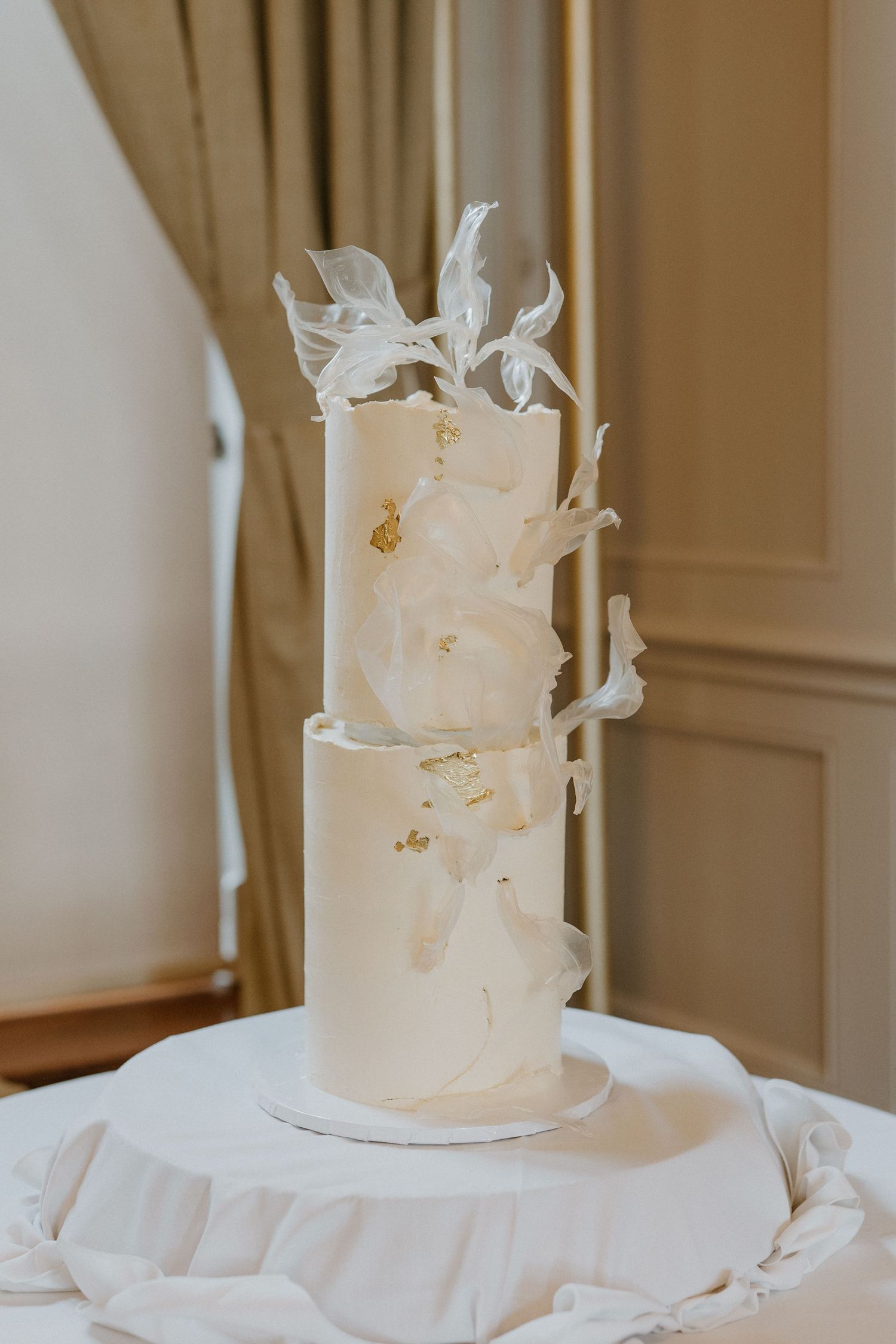 Two-tier white wedding cake decorated with sculpted sugar flowers and gold leaf accents.