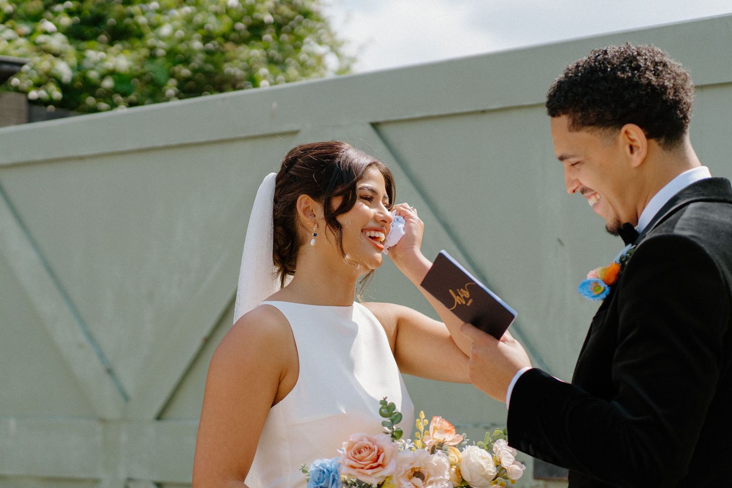 Groom in black tuxedo gently wipes tears from his bride's cheek during their wedding ceremony.
