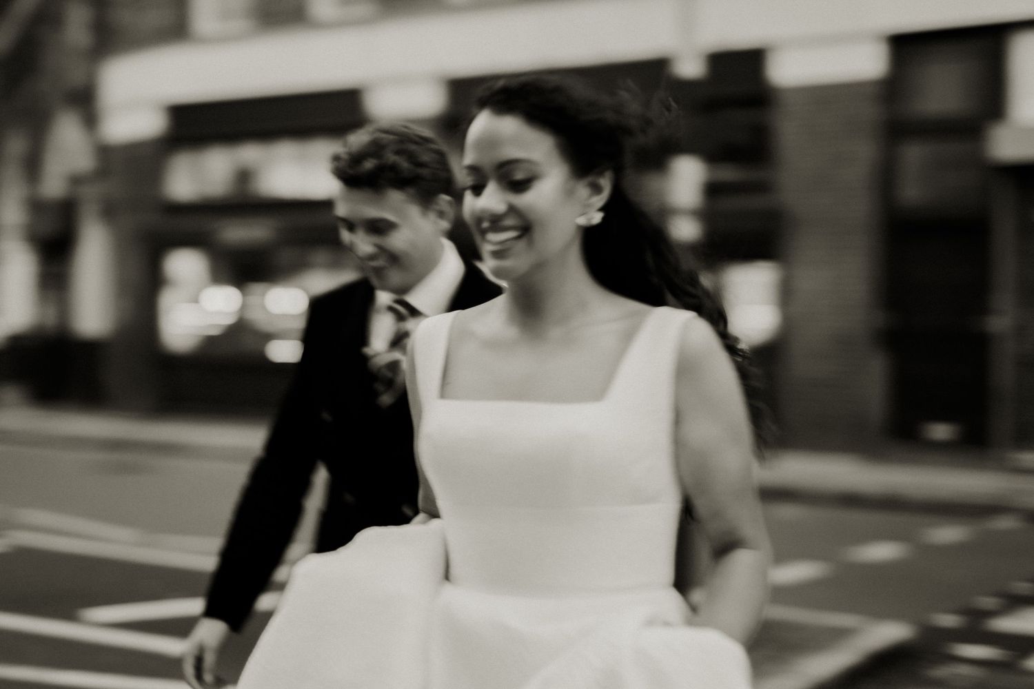 Black and white photo of smiling bride in white dress walking on city street.