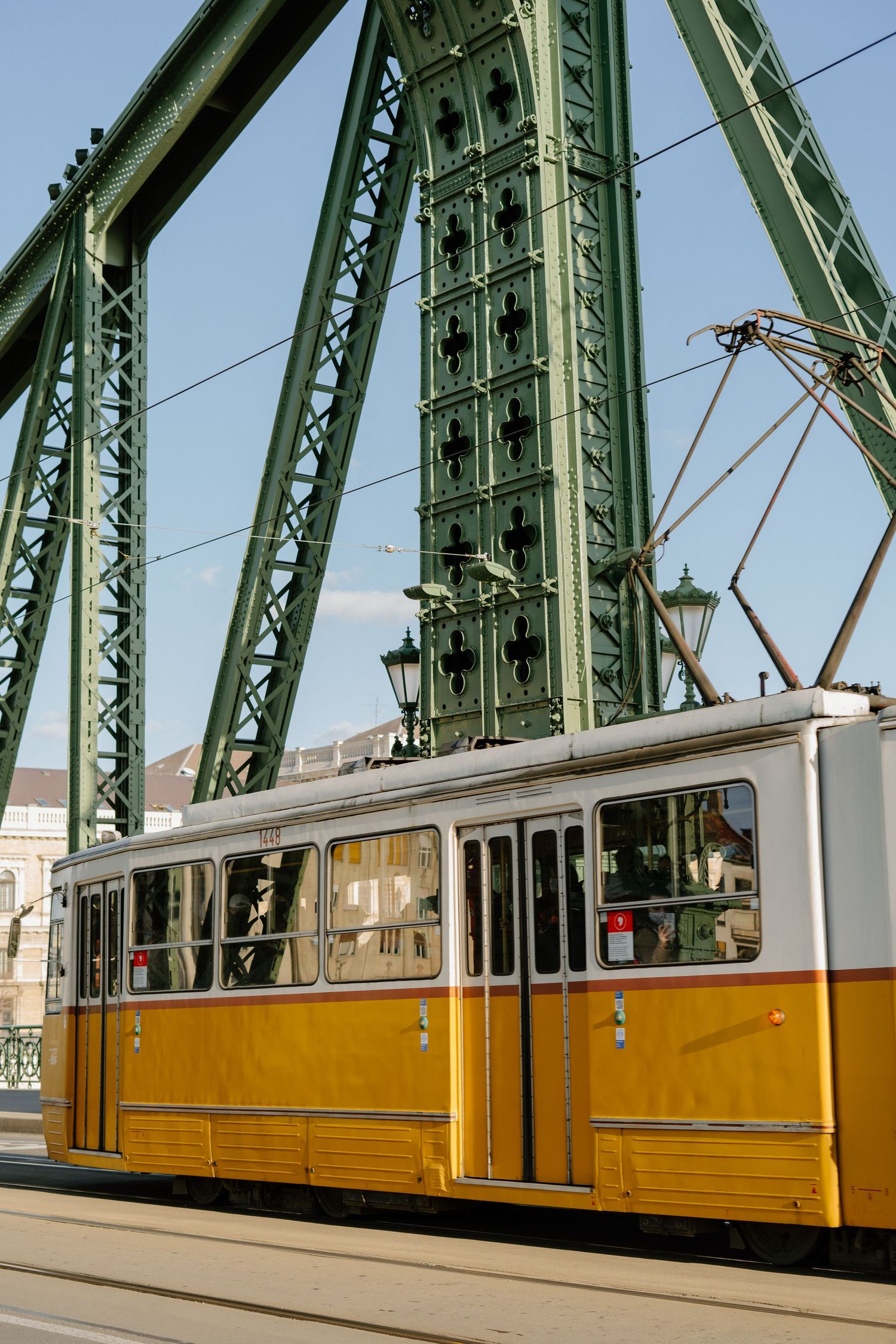 A vintage yellow and white tram passes under the green steel beams of Budapest's Liberty Bridge on a sunny day.