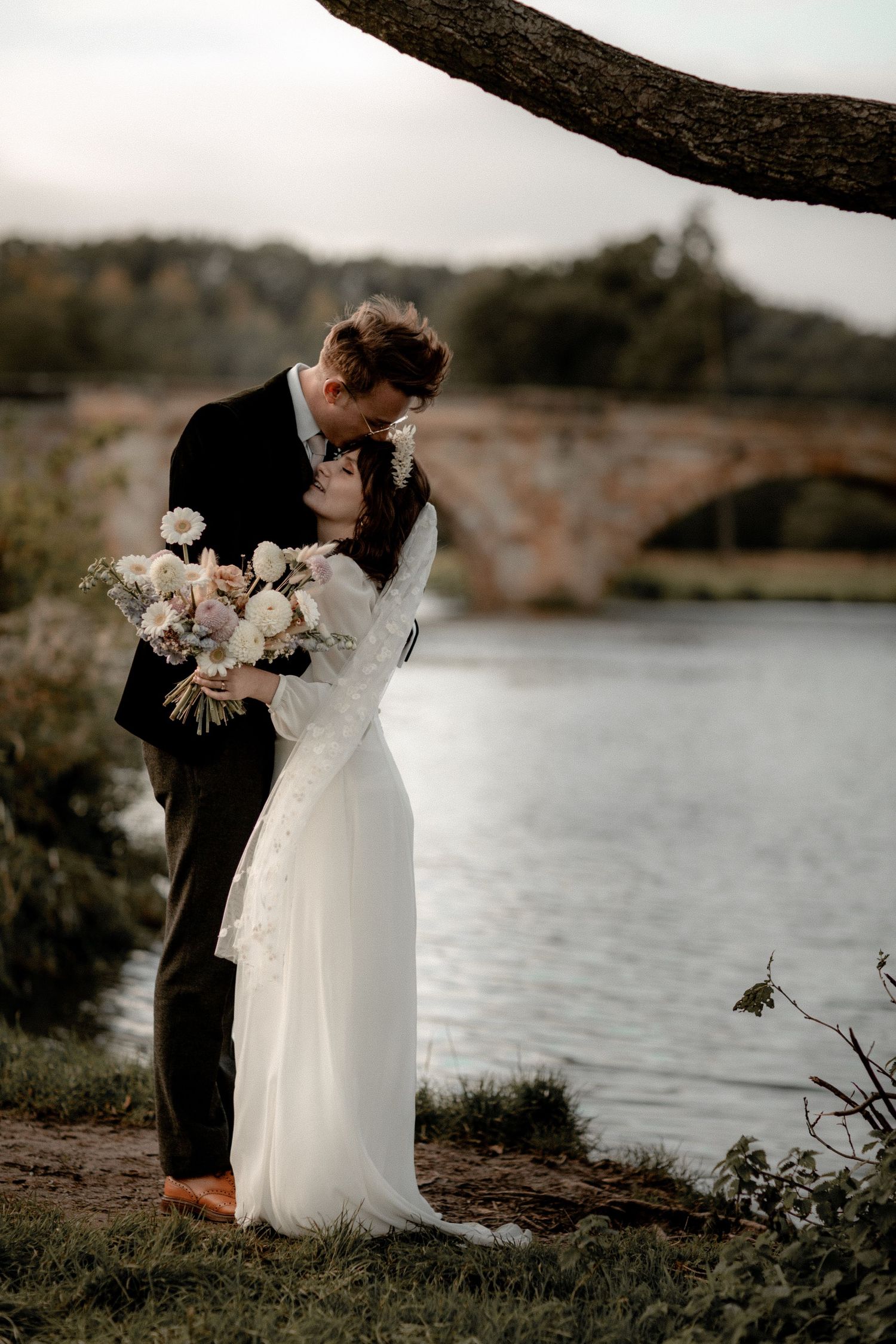 A romantic wedding portrait by a stone bridge over water in moody evening light.