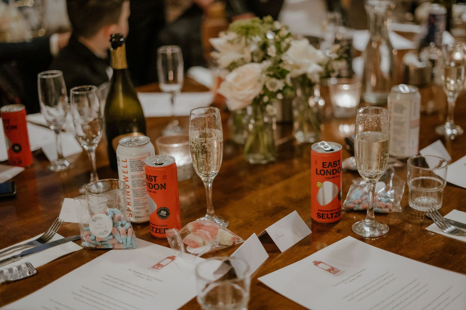 Elegant wedding reception table with champagne flutes, floral centerpiece, candles, and beverage cans on a wooden table.