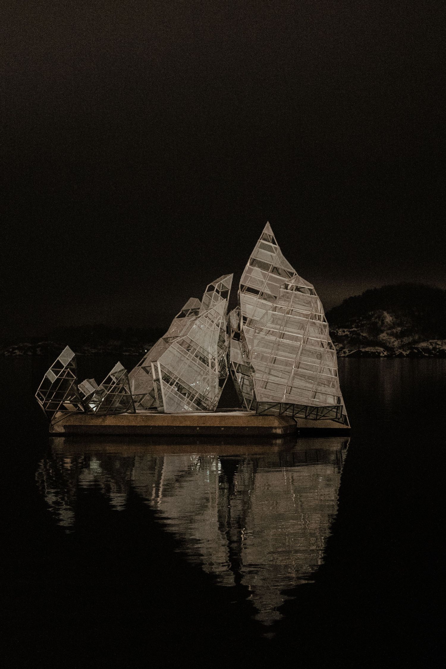 Illuminated geometric glass sculptures float on dark water creating mirror-like reflections at night.