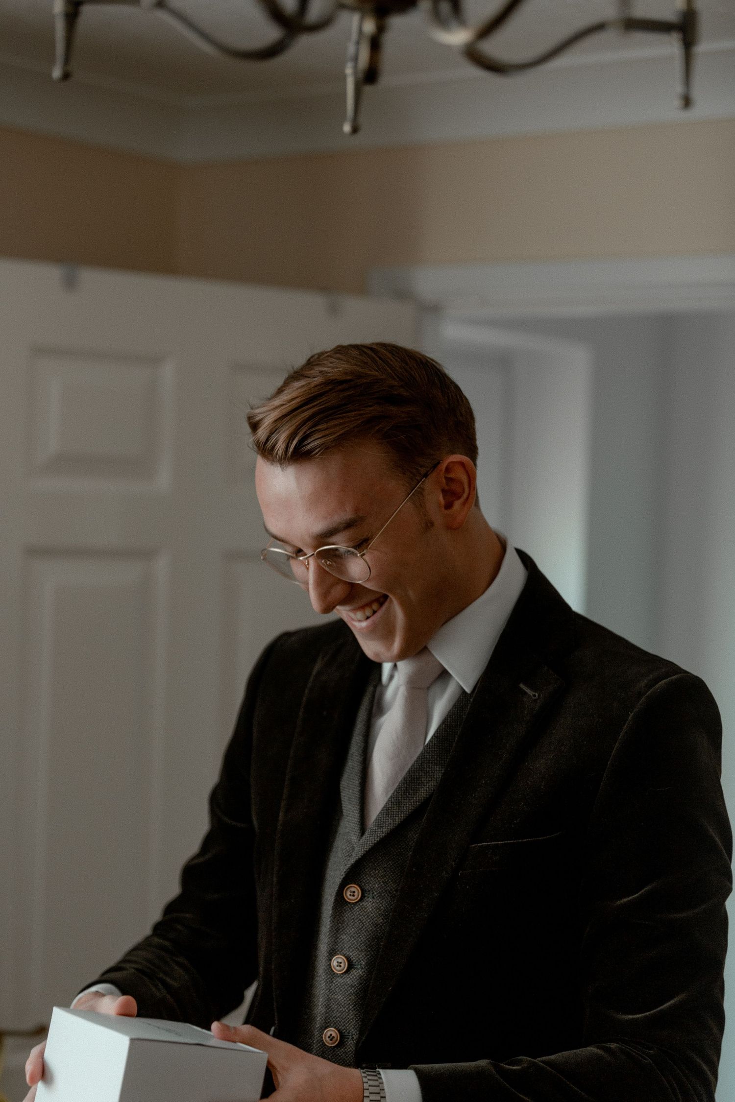 A person in a dark suit and glasses smiles while opening a gift box indoors.