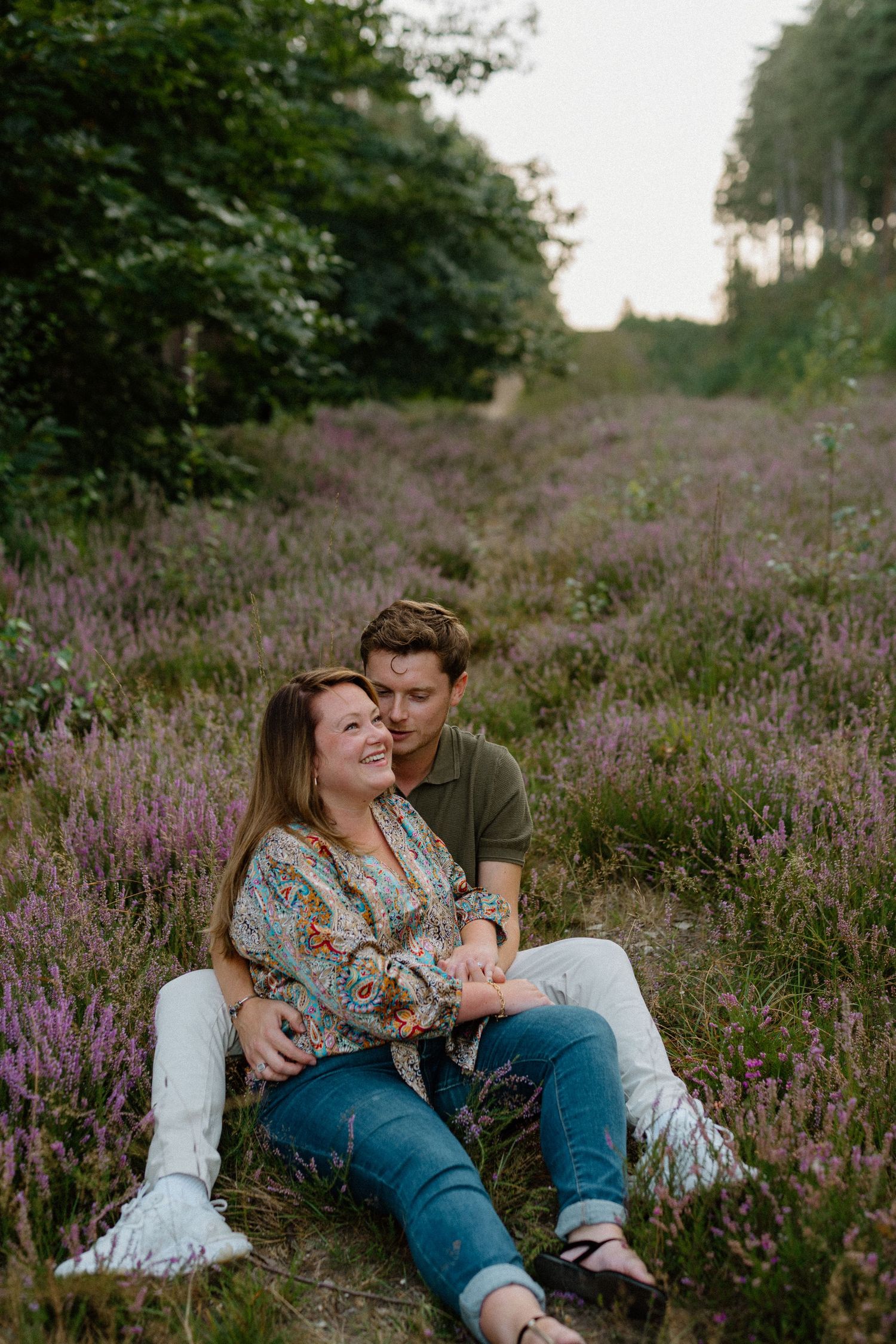 Couple sitting together in a blooming purple heather field at sunset surrounded by trees.