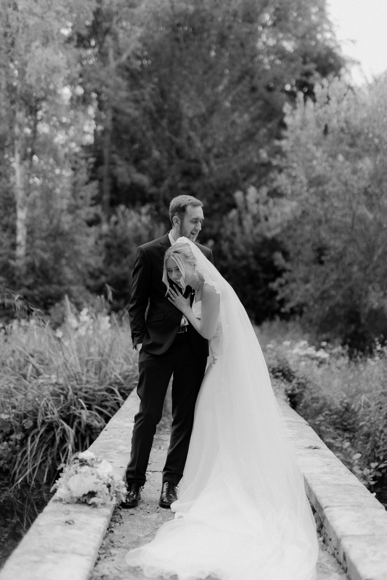 Black and white photo of groom embracing bride in flowing wedding dress on stone path surrounded by lush greenery.