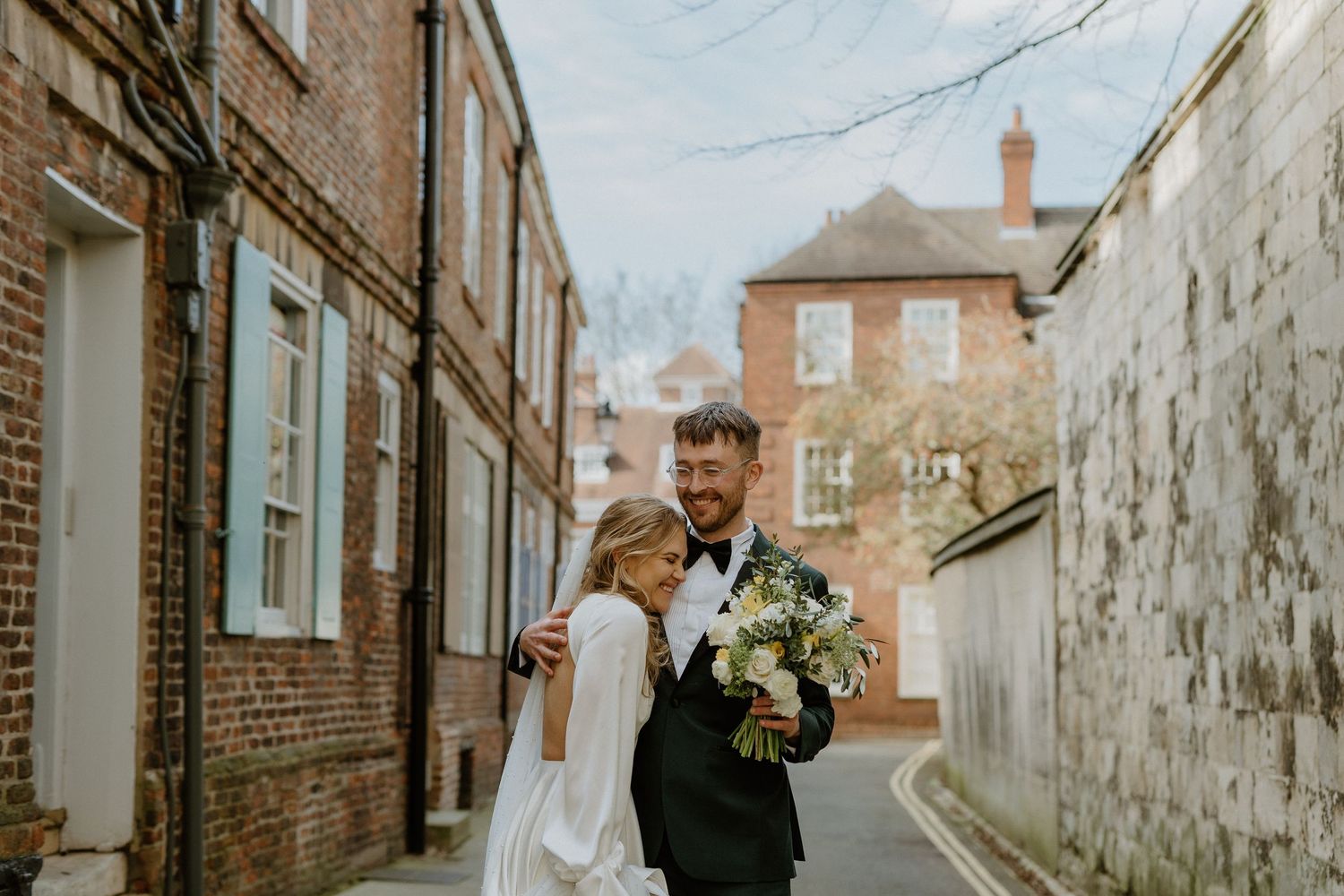 Bride and groom walking together in a charming brick alley, groom holding a white bouquet, both smiling on their wedding day.