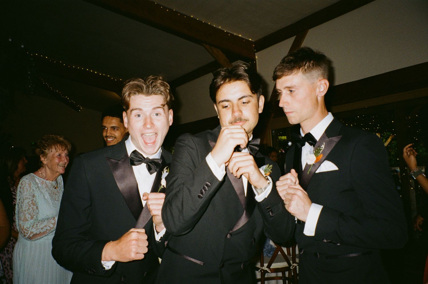 Three groomsmen in tuxedos singing into a microphone at a wedding reception.