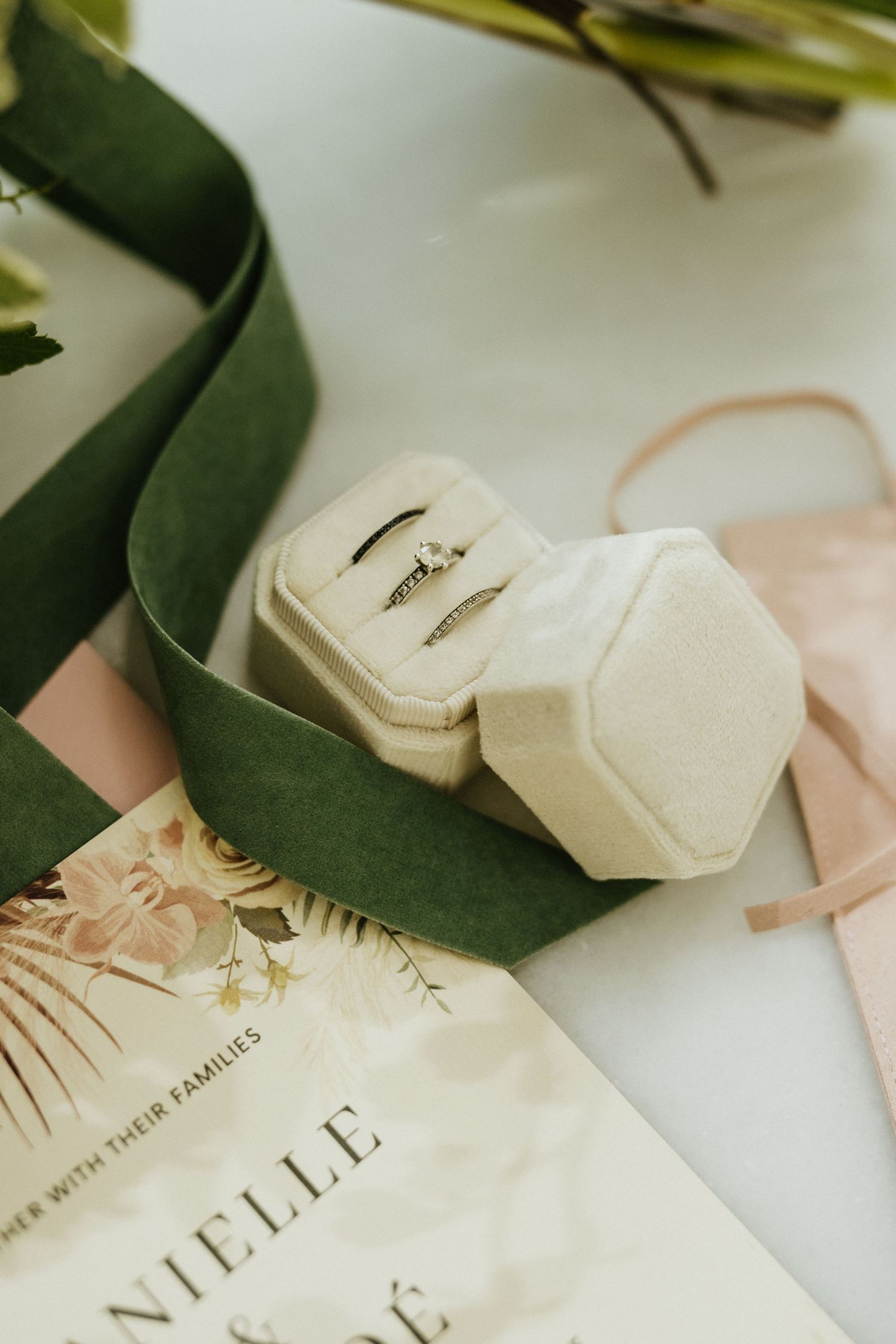 Elegant wedding rings displayed in a cream velvet hexagon box with green ribbon and floral invitation.