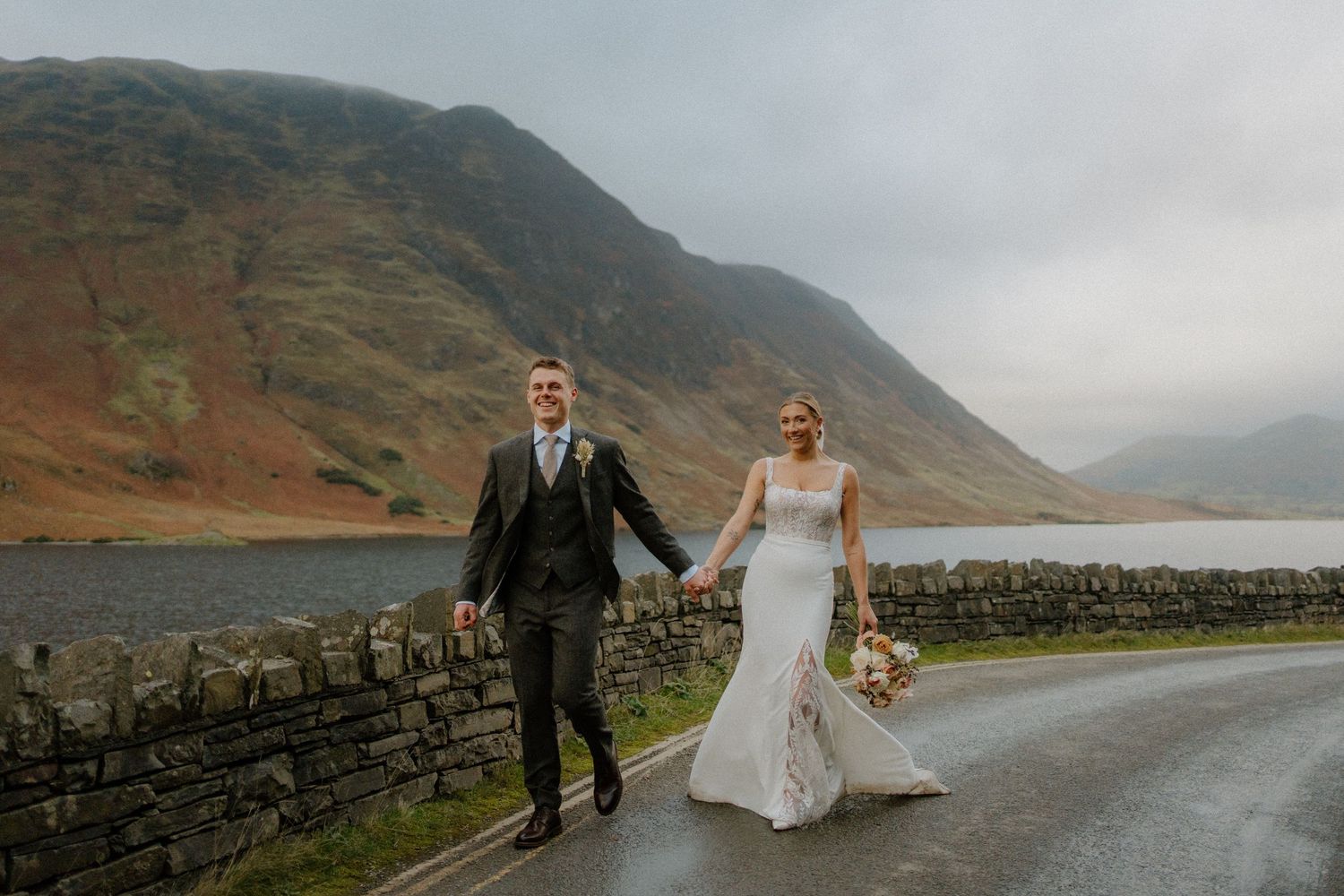 Bride and groom walk hand-in-hand along a rainy lakeside road with dramatic misty mountains in the Lake District.