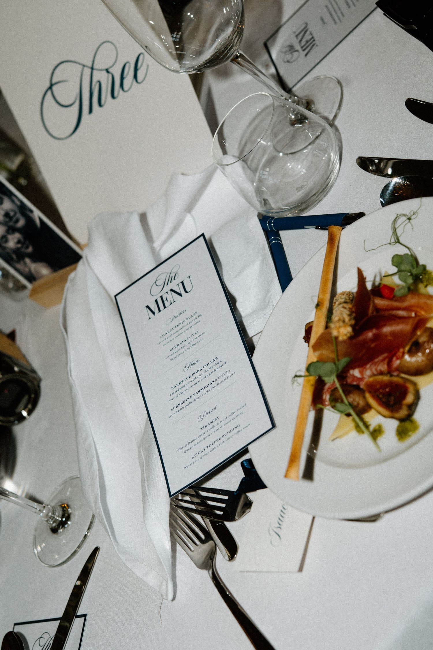 Elegant wedding reception table setting with menu card, white napkin, crystal glassware, and a plated gourmet starter at Table Three.