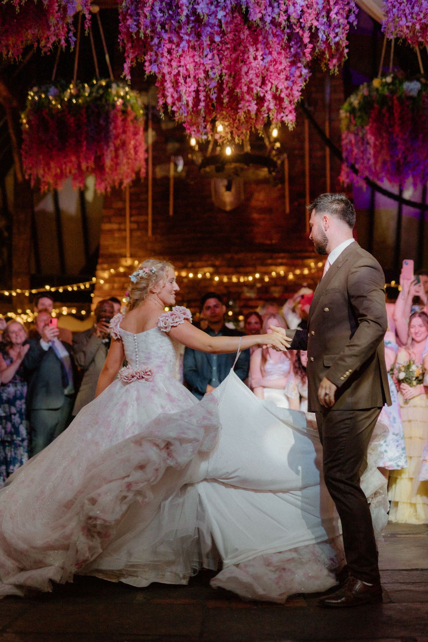 Bride and groom dance beneath cascading purple flowers with fairy lights and cheering guests in barn.