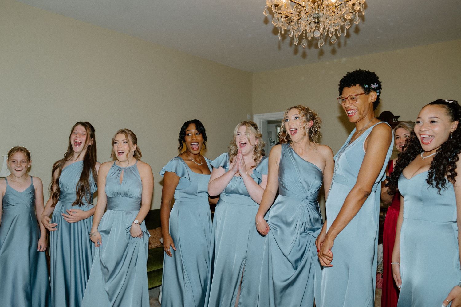 Excited bridesmaids in blue dresses reacting with joy beneath a crystal chandelier.
