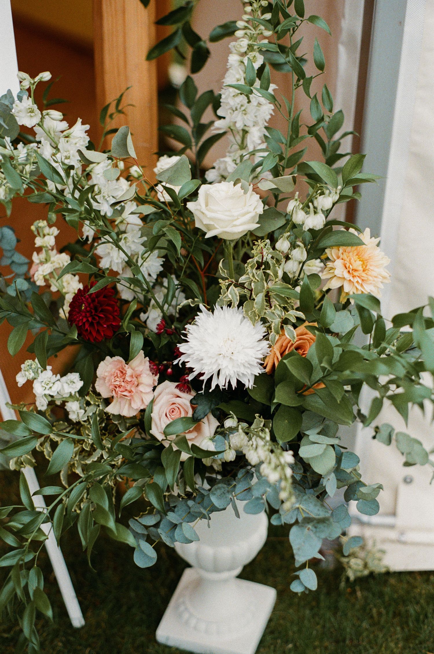 Lush floral arrangement with white, pink, and burgundy blooms in white urn at outdoor wedding ceremony.