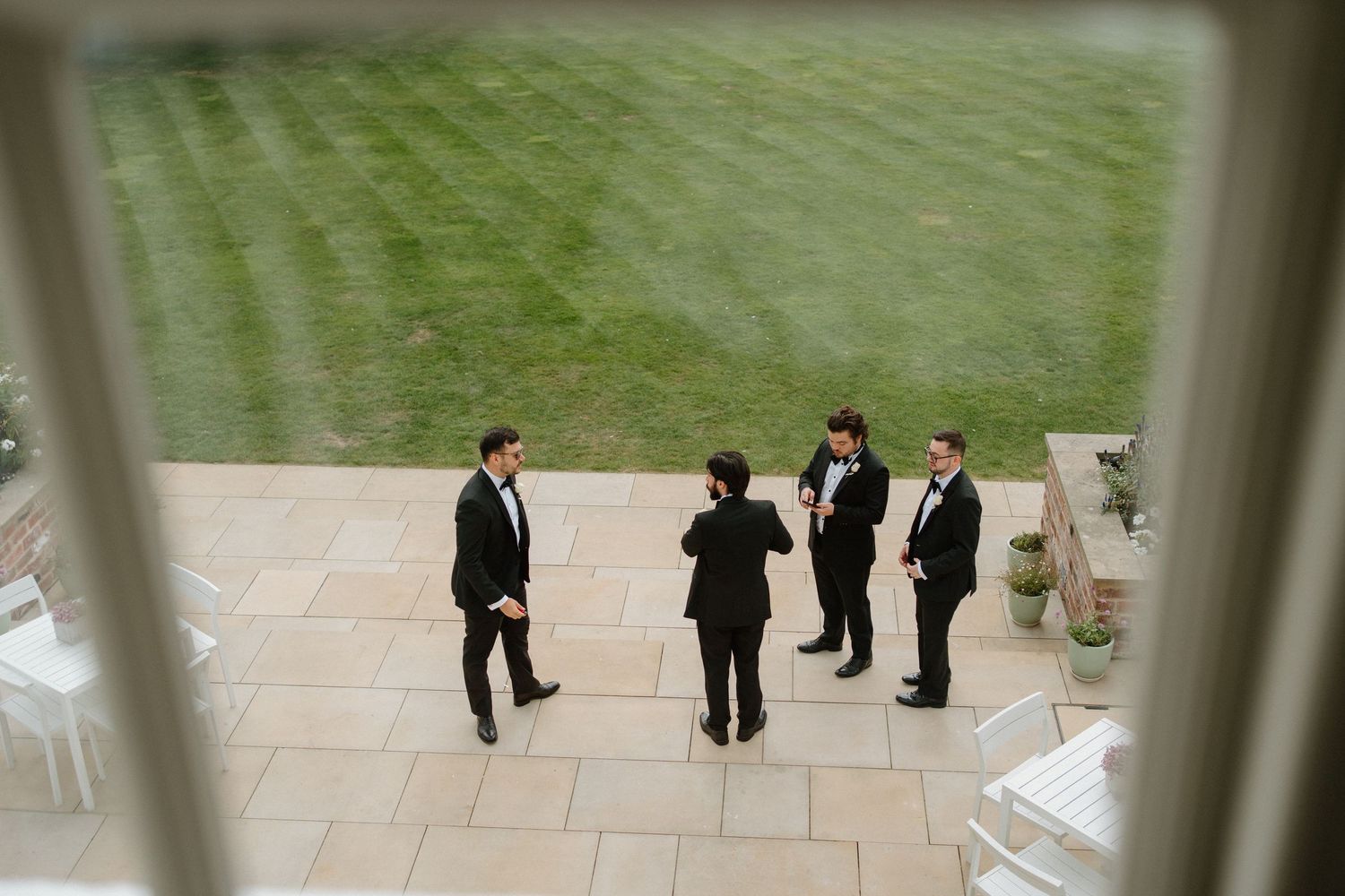 Aerial view of groomsmen in black tuxedos chatting on stone patio beside manicured lawn.