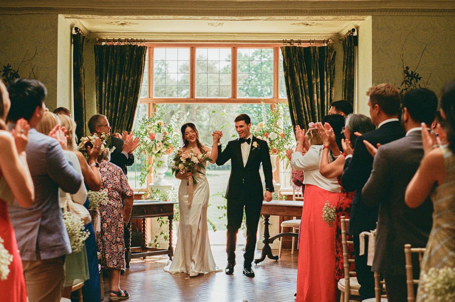 Bride and groom exit ceremony as guests applaud in elegant floral-decorated indoor wedding venue.