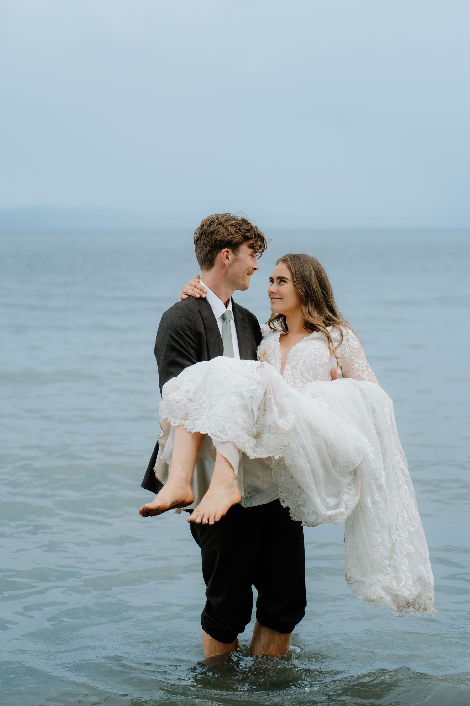 Groom carries smiling bride in white lace dress while standing in shallow ocean water.