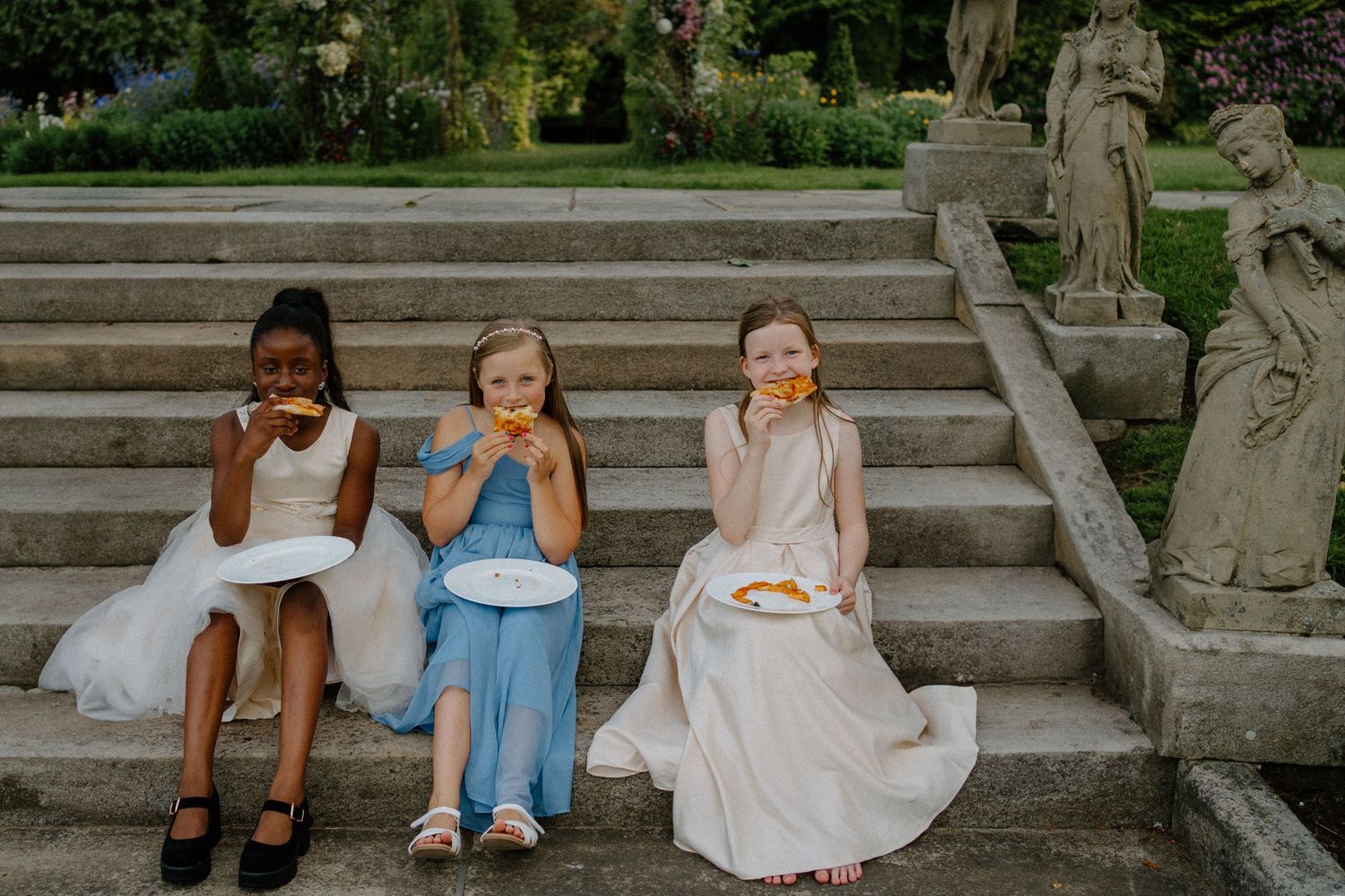 Three flower girls in elegant dresses sitting on stone steps eating pizza at a wedding reception outdoors.