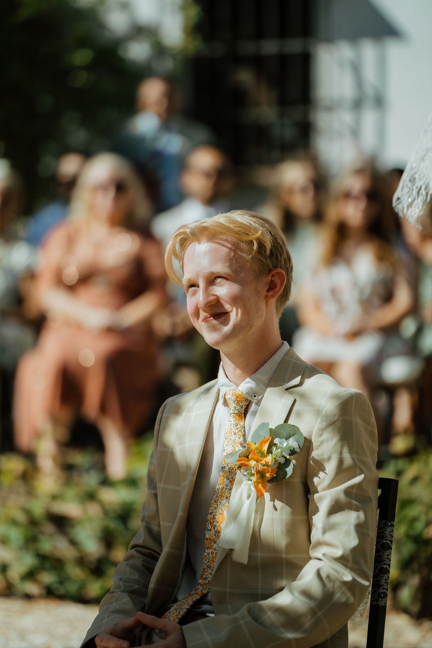 Smiling groom in a plaid suit with floral boutonniere sits at the altar during an outdoor wedding ceremony.