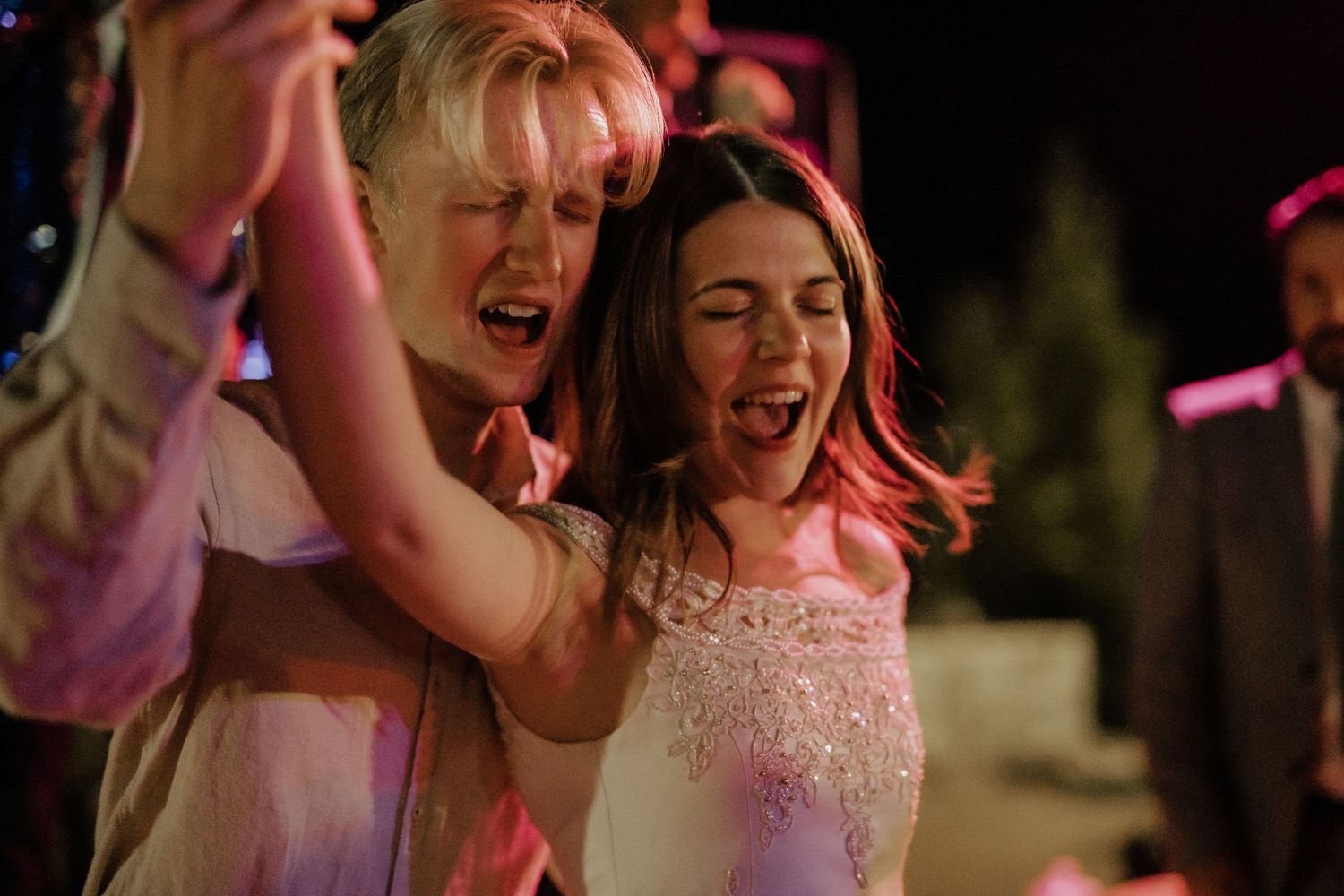Two women joyfully singing and dancing together at a wedding reception under pink stage lights at night.